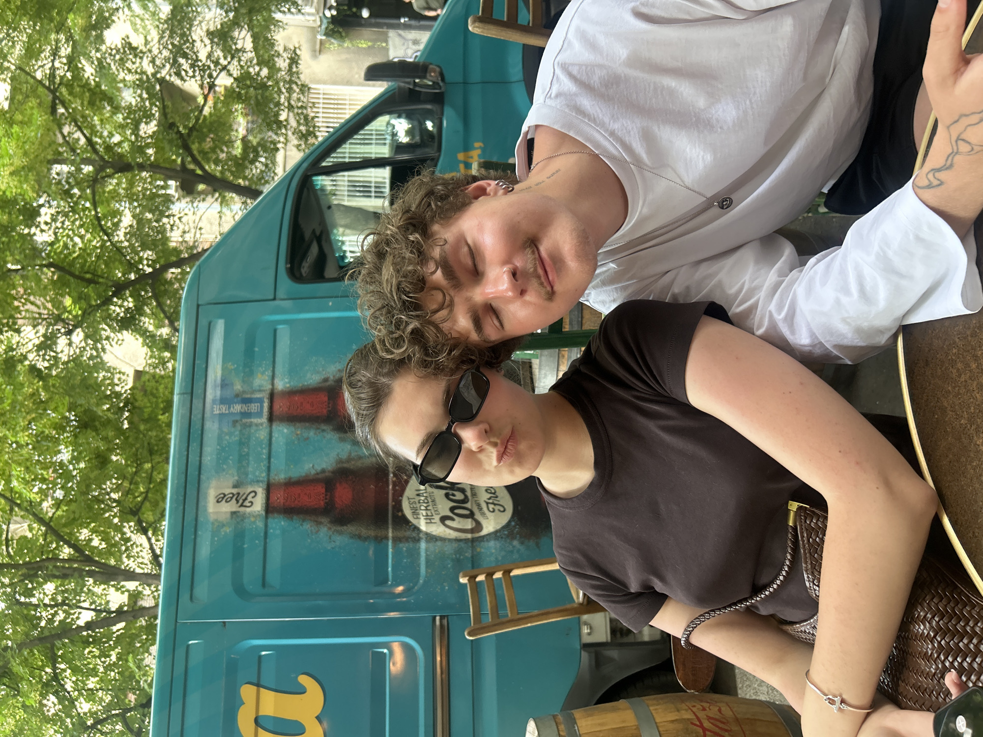 Two people are sitting at an outdoor table. A woman with sunglasses and a dark t-shirt is seated at the table. A man with curly hair and a white shirt is leaning towards her. Behind them is a turquoise van with Coca-Cola graphics and the word "Free" on the side. Trees and buildings are visible in the background. The setting appears to be an outdoor cafe in an urban environment. Given the "Free" Coca-Cola branding on the van and the architectural style, the location is likely in Belgrade, Serbia.