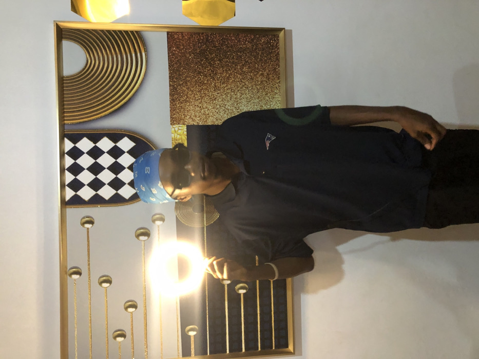 This image, captured in Tema, Ghana, features a single male individual standing indoors. The subject, appearing to be a young adult, is centrally positioned and faces the viewer. He wears a blue bandana with white patterns, dark sunglasses, a navy blue short-sleeved polo shirt bearing the New England Patriots logo on the left chest, and dark trousers. A white bracelet is present on his left wrist. His right hand holds a brightly illuminated, circular light source, extended forward and slightly upwards, which emits an intense glow and creates a halo effect. Behind the individual, a large, framed piece of wall art is visible, comprising multiple abstract sections: a left portion with gold horizontal lines and spherical embellishments on a grey background, a central upper section with a black and white checkered shield design, a right upper section displaying concentric gold curved lines, and a lower right section featuring a textured, golden-brown surface. To the far right of the frame, two partially visible, yellow-gold, geometric light fixtures or decorative elements are present, also emitting light. The wall serving as the background is light-colored.
