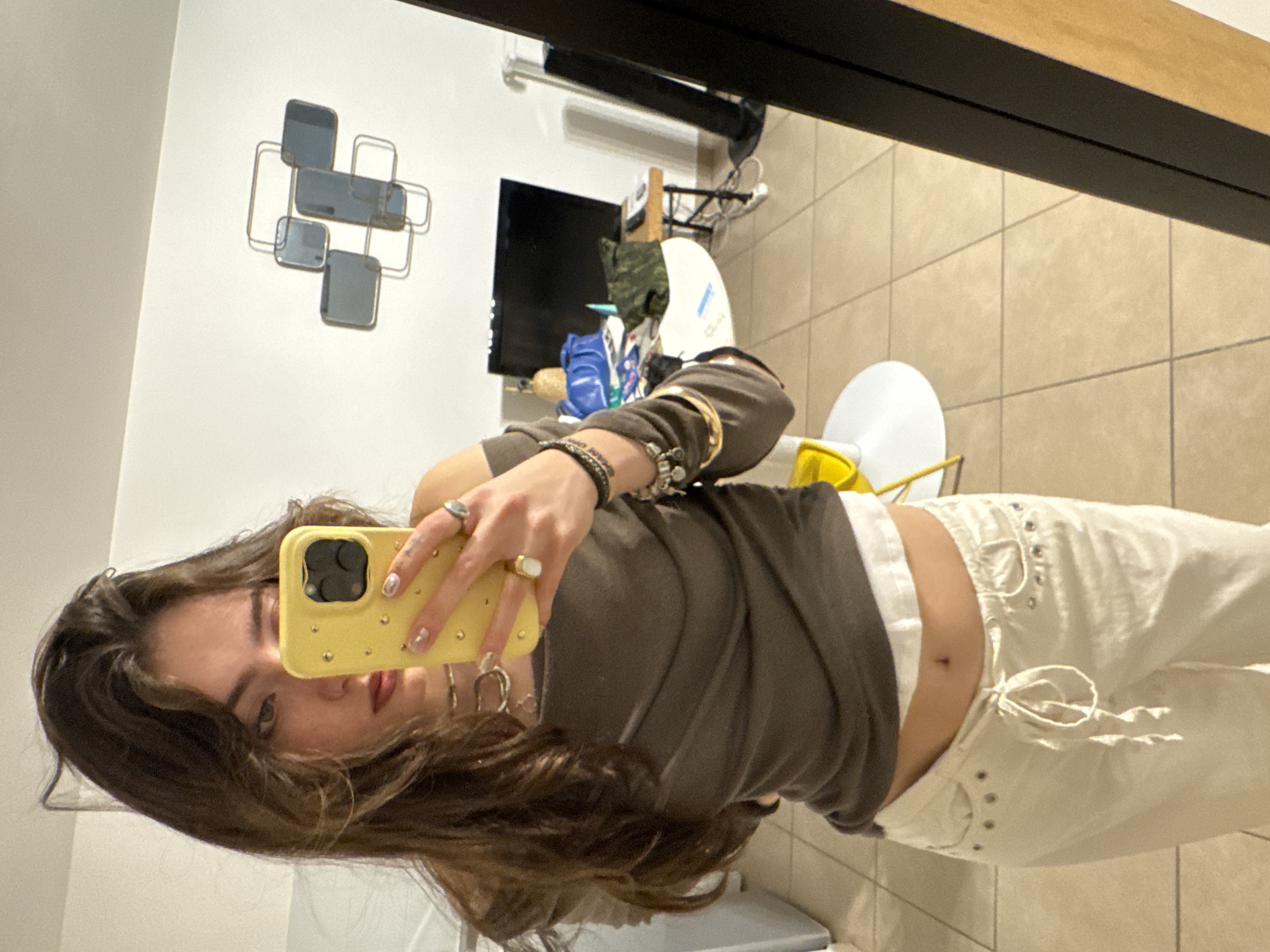 A young woman takes a mirror selfie in an interior space. She is wearing a brown long-sleeved shirt and white pants. The background includes a wall decoration consisting of geometric shapes, a television, and various objects on a shelf or counter. The floor is tiled. The location is not identifiable as Saint-Ouen-sur-Seine, France from this image.