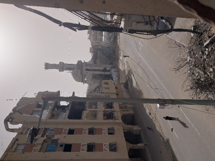 An aerial view shows extensive war damage. Buildings are shelled, particularly a large apartment block. A mosque with a minaret stands in the distance. Debris is scattered everywhere. A lone figure walks down an empty road. Wires and parts of a collapsed building are visible in the foreground, likely from where the photo was taken.