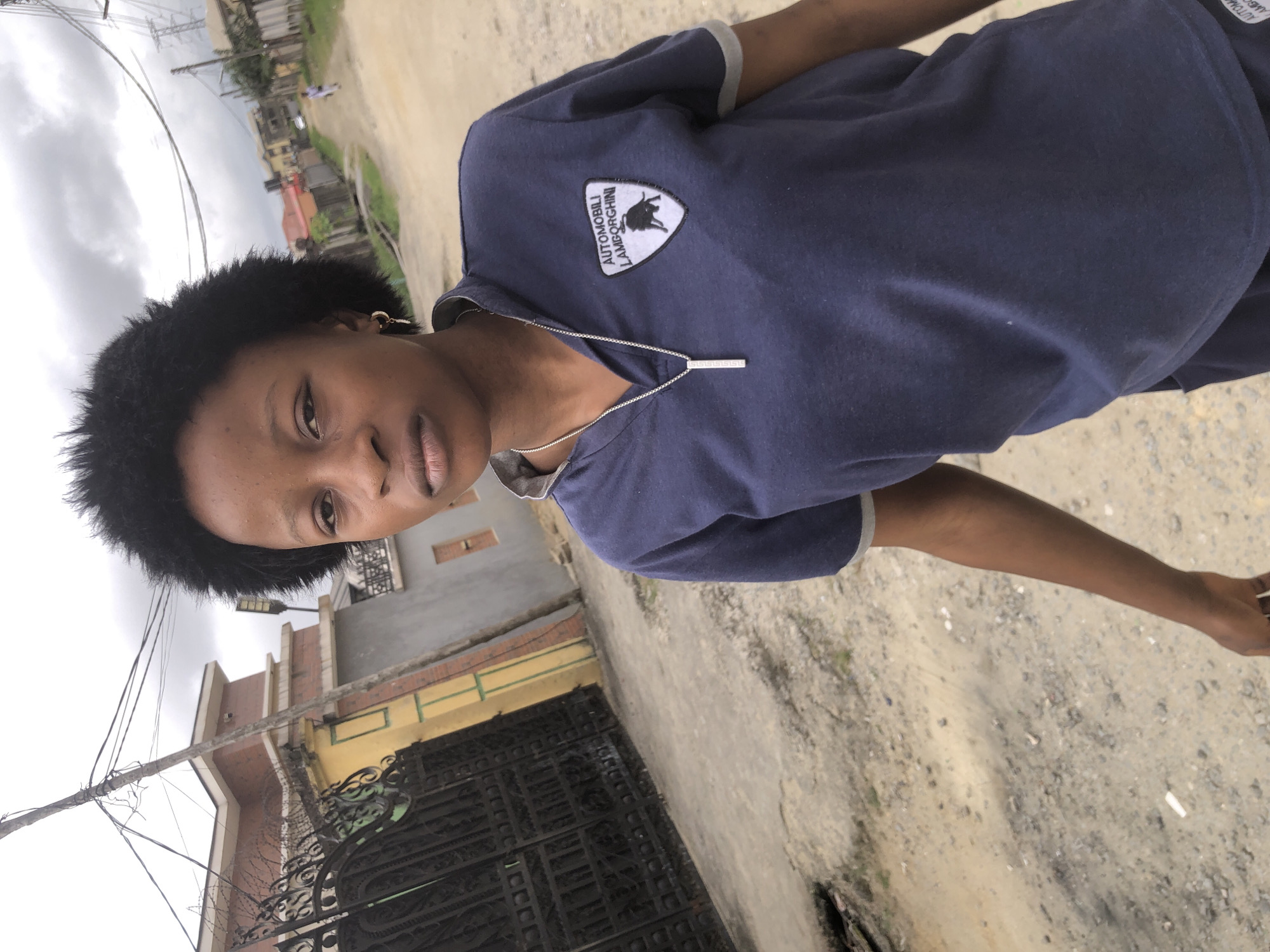 A young woman with dark, curly hair and a dark blue shirt with a Lamborghini logo stands on a dirt road in what appears to be a residential area of Ado, Nigeria. The woman is looking directly at the camera with a neutral expression. The road is unpaved and has loose gravel and dirt. To the left of the woman is a large black metal gate, which appears to be the entrance to a property. Behind the gate, there is a building with a yellow and brown facade, and a red-tiled roof. A utility pole with numerous wires stands near the gate. In the background, other buildings and houses are visible, as well as more utility poles and wires. The sky is overcast. The overall scene depicts a street in a Nigerian city with typical infrastructure and residential architecture.