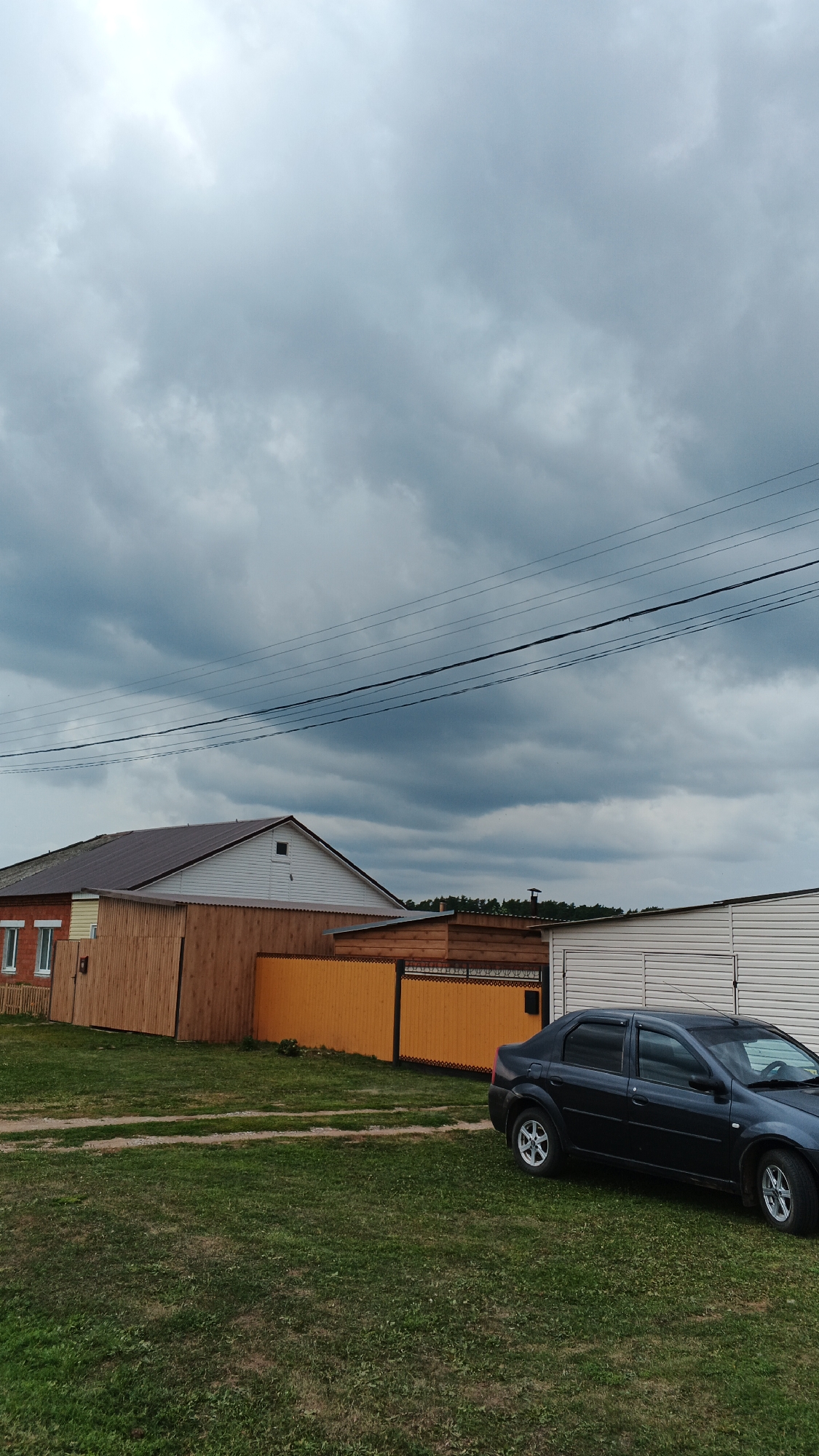 The image depicts a residential scene on a cloudy day in the village of Podgornoye, Russia. In the foreground, a dark-colored sedan is parked on a grassy area with a dirt path. Behind the car and to the left, there are several structures, including a house with a brown roof and white siding, adjacent to a wooden fence and an orange metal fence. Further to the right, a white metal building with a corrugated exterior is visible. Overhead, power lines crisscross the sky. The overall environment appears to be a rural or semi-rural setting with residential buildings and vegetation.