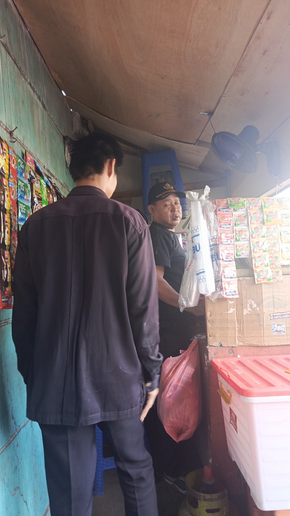 Two men are standing inside a small, open-air stall. The man on the left is facing away from the camera, wearing a dark blue button-up shirt and dark pants. The man on the right is facing the camera, wearing a black t-shirt and a black baseball cap with gold trim. He is holding a stack of clear plastic cups. The stall is stocked with various packaged goods, including tea and what appear to be snacks or instant noodles. A blue plastic stool is visible behind the man on the left, and a red and white plastic container is on a shelf to the right. A small fan is mounted on the ceiling. The stall appears to be constructed from wood and plywood. The environment suggests an informal marketplace or a small convenience store. The scene is likely located in Tangerang, Indonesia, given the context provided.