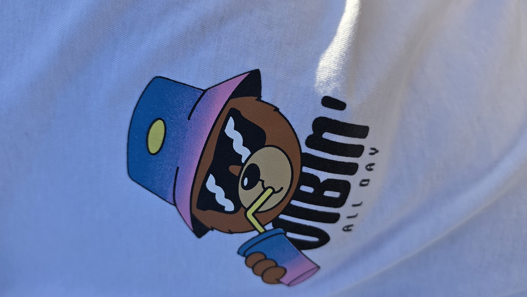 The image shows a close-up of a white t-shirt with a graphic print. The graphic features a cartoon bear wearing a bucket hat and sunglasses, holding a cup with a straw. The text "VIBIN' ALL DAY" is printed below the bear. The t-shirt appears to be new or in good condition, with a clean white fabric. The lighting suggests an outdoor setting, possibly on a sunny day, as indicated by the shadow cast on the fabric. The image does not provide any information about its location in Jūrmala, Latvia.