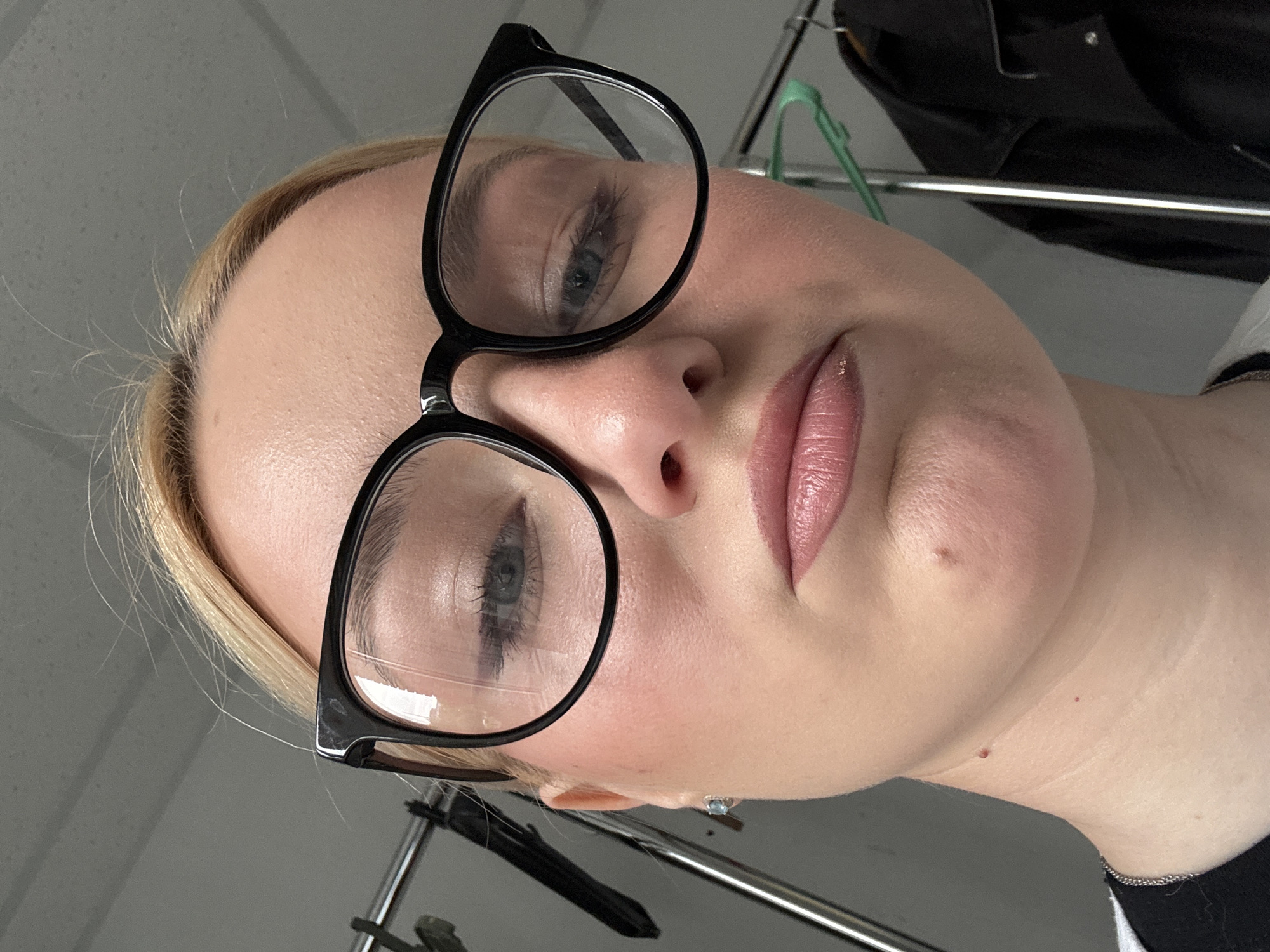 A close-up, low-angle selfie of a fair-skinned woman with blonde hair pulled back is captured. She is wearing black-rimmed glasses and has a pale pink lipstick on. Her expression is neutral, with her mouth slightly downturned. A small mole is visible on her chin, and another on her neck, below a faint scar. She is wearing a black shirt with a silver chain necklace. The background is blurred, showing a metal clothing rack with garments hanging on it, and a light grey wall. There are no identifiable landmarks or elements that place this image in Sinyavino, Russia.