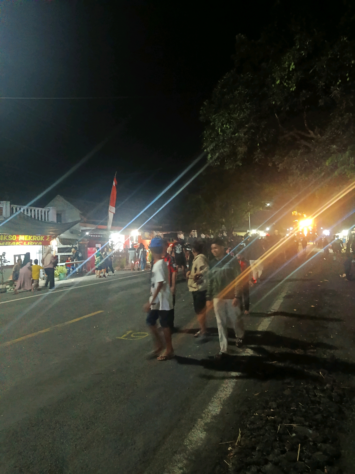 The image depicts a nighttime scene on a street in Pasirian, Indonesia, likely near a market or gathering area. Several individuals are visible walking on the asphalt road, with buildings and stalls lining the street. The lighting appears to be artificial, coming from various sources, including streetlights and possibly the stalls themselves, creating bright lens flares across the image. A national flag, red and white, is prominently displayed on a pole. The ground is dark asphalt with yellow markings. In the foreground, the legs and feet of a person are visible, wearing flip-flops. The overall atmosphere suggests a bustling evening event.