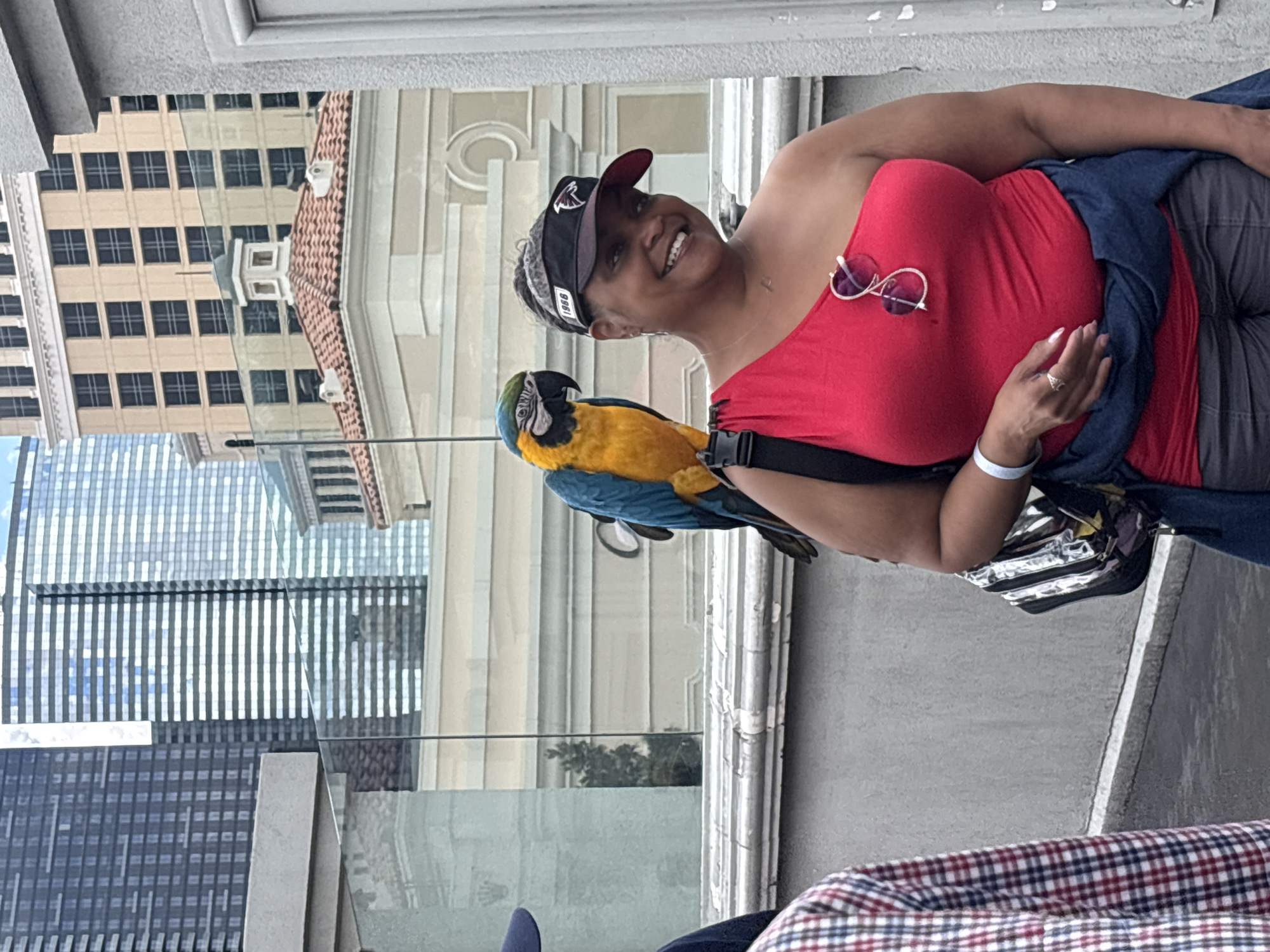 A medium shot depicts a smiling African American woman with a parrot perched on her shoulder, taken outdoors. The woman is wearing a red, off-the-shoulder top, a dark visor with the letter “A,” and carries sunglasses. Her left hand is raised slightly with a ring visible on her finger. A blue and gold macaw is perched on her left shoulder, attached by a black strap. The urban background suggests a balcony setting in Paradise, Nevada, United States of America. Buildings of varying heights and styles are visible across the street, including a modern glass building reflecting the sky, and a beige building with multiple windows and a red-tiled roof. The foreground includes a small portion of plaid fabric.