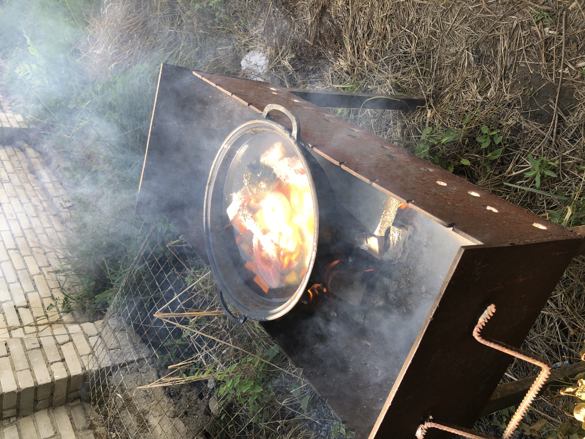 A Dutch oven filled with food and liquid is placed on a rustic metal grill over an open fire. Smoke billows from the fire, obscuring part of the grill and the surrounding vegetation. The grill is positioned outdoors on dry, brown grass and weeds, with a rough, light-colored brick wall and a wire fence visible in the background. The overall scene suggests an outdoor cooking activity, possibly in a backyard or a more rural setting. The image does not provide any specific visual cues to pinpoint a location within the city of Lviv, Ukraine.