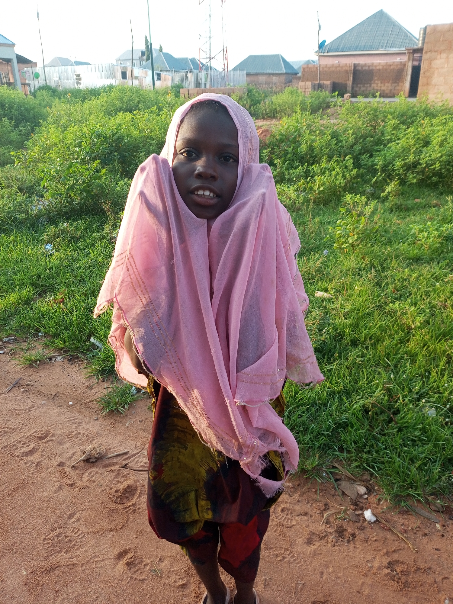 A young Black child, adorned in a light pink headscarf partially covering their face and chest, stands outdoors on a sandy path. Their gaze is directed towards the camera. The child wears a dark skirt with a vibrant, abstract pattern of green, yellow, and red. Behind the child, a landscape of lush green grass and scattered shrubbery stretches towards a backdrop of buildings with corrugated iron roofs and brick construction. The sky above is overcast. The scene appears to be a rural or semi-urban area within Tempure, Nigeria, characterized by its natural vegetation and developing infrastructure.