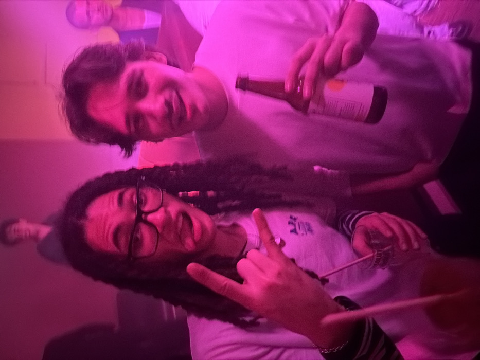 Two people pose closely together in a dimly lit room with pinkish-purple lighting. The person in front wears glasses and a striped-sleeved shirt, making a rock-on hand gesture. The person behind holds a bottle. Faces of other people are partially visible in the background, one in the upper left and another further back near the bottom. The overall image has a casual, candid feel, likely taken at a social gathering or party.