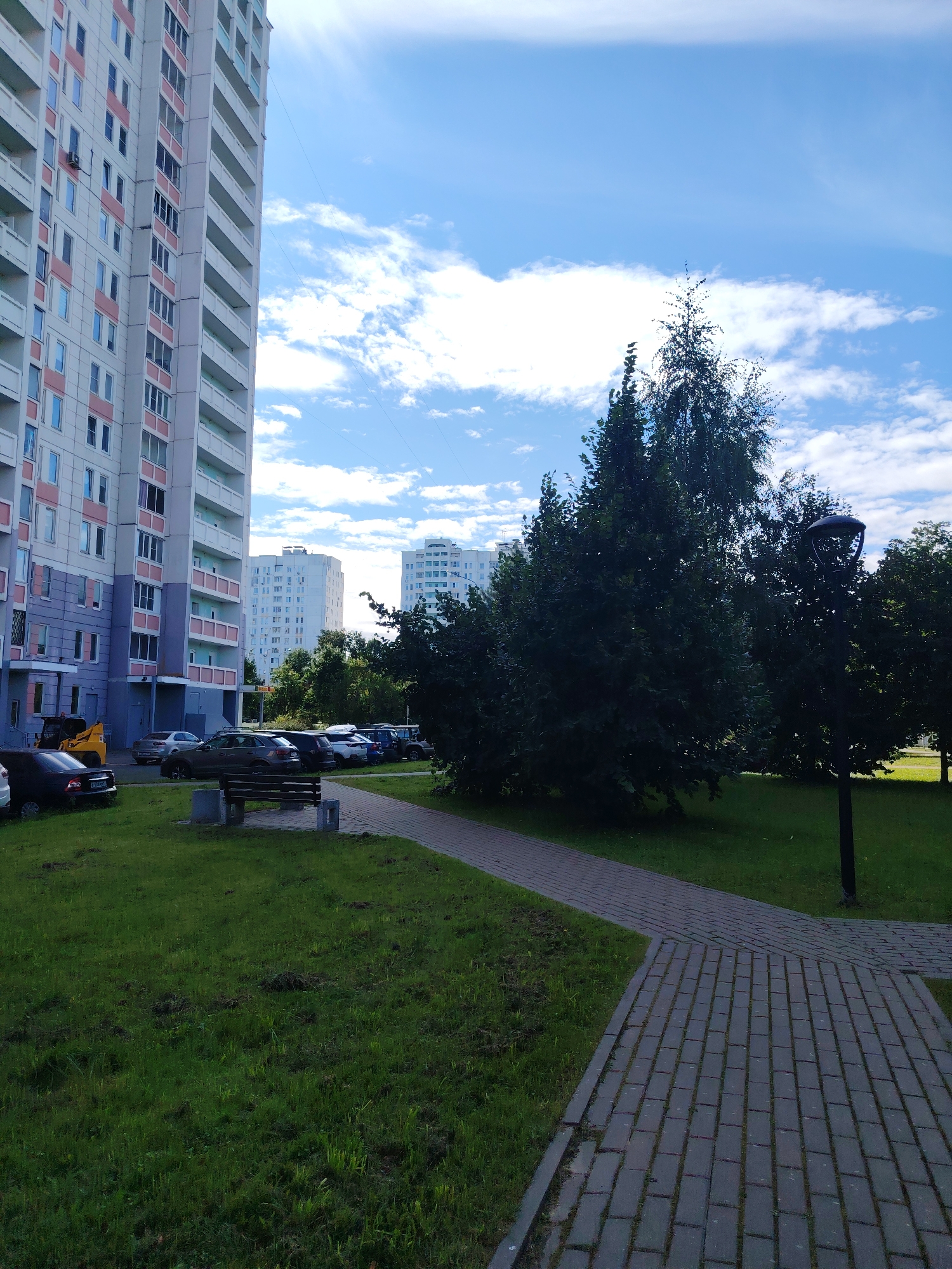 A paved pathway curves through a grassy area in front of a tall, multi-story residential building in Zelenograd, Russia. The building, with its light-colored facade and pink accents, dominates the left side of the frame. To the right of the pathway, a dense stand of trees provides shade. Several cars are parked in a lot adjacent to the building. A wooden bench with concrete supports sits on the grass near the pathway. The sky above is a vibrant blue, dotted with fluffy white clouds. In the background, two more residential buildings can be seen. The scene is illuminated by bright daylight.