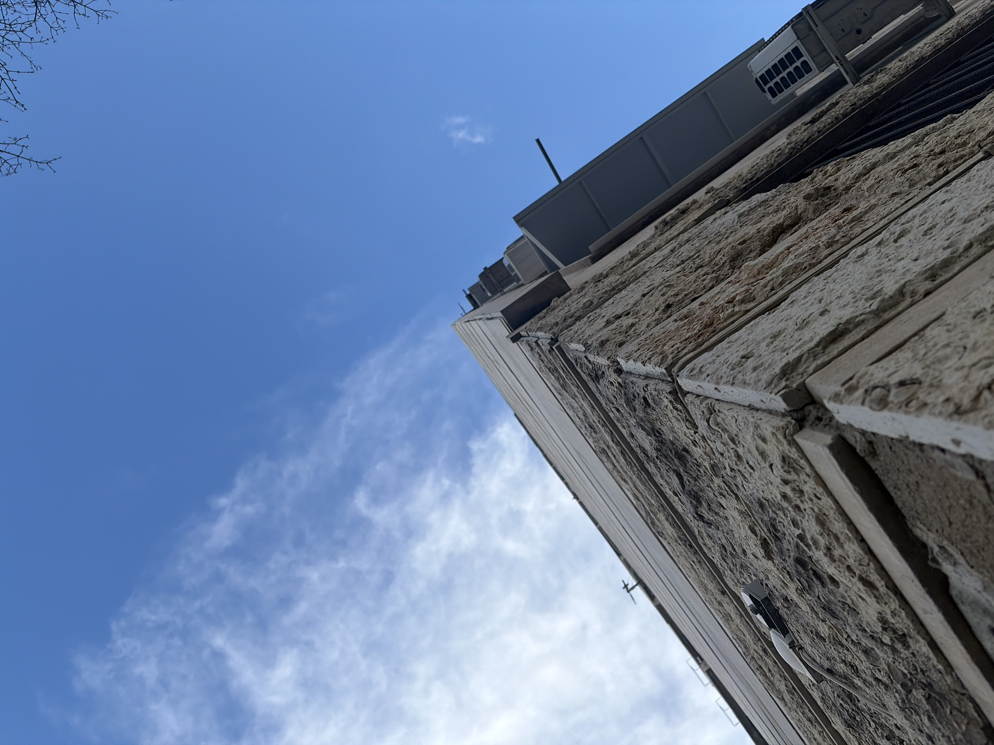 The image shows a low-angle view of a building, possibly in Sofia, Bulgaria, characterized by its stone facade. The sky is partially visible, with scattered clouds. The building appears to have a gray, possibly metallic, structure on top, which may house mechanical equipment. A small part of a tree branch is visible in the top-left corner.