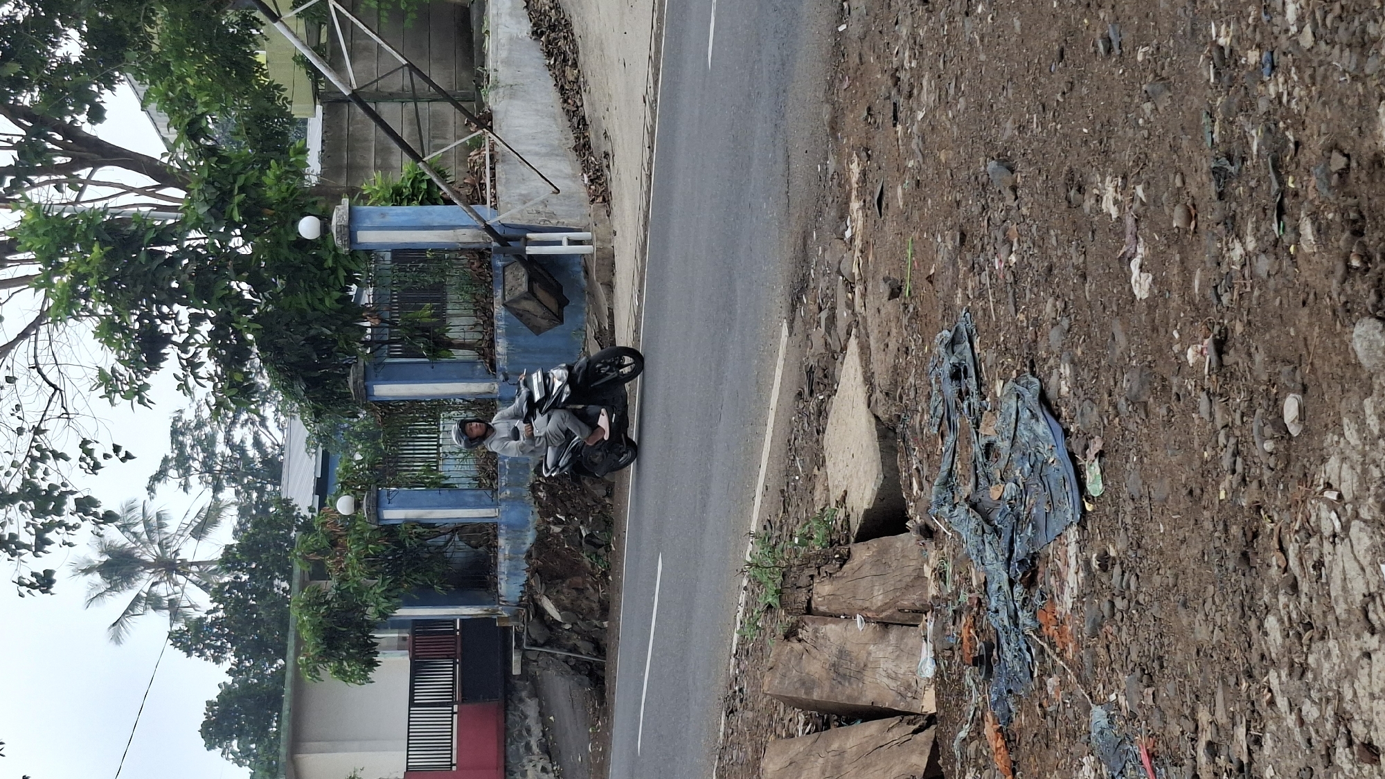 A person wearing a grey hoodie and helmet rides a black motorcycle along a paved road in Cilaku, Indonesia. The rider is in motion, facing right, with their feet on the footrests. The motorcycle is a scooter-type, with a visible exhaust pipe and rear suspension. The road is asphalt with a white line dividing the lanes. To the left of the road, there is a red and white building with a black fence. Beyond that, a blue and white striped fence encloses an area with trees and vegetation. To the right of the road, a concrete wall and some metal structures are visible, along with fallen leaves and dirt. The foreground of the image shows a rough, dirt path covered with debris, including pieces of wood, torn blue tarp, and various other discarded items. The environment appears to be a roadside area, likely in a semi-rural or developing urban setting, given the mix of structures, vegetation, and accumulated refuse. The sky is overcast, suggesting a cloudy day.