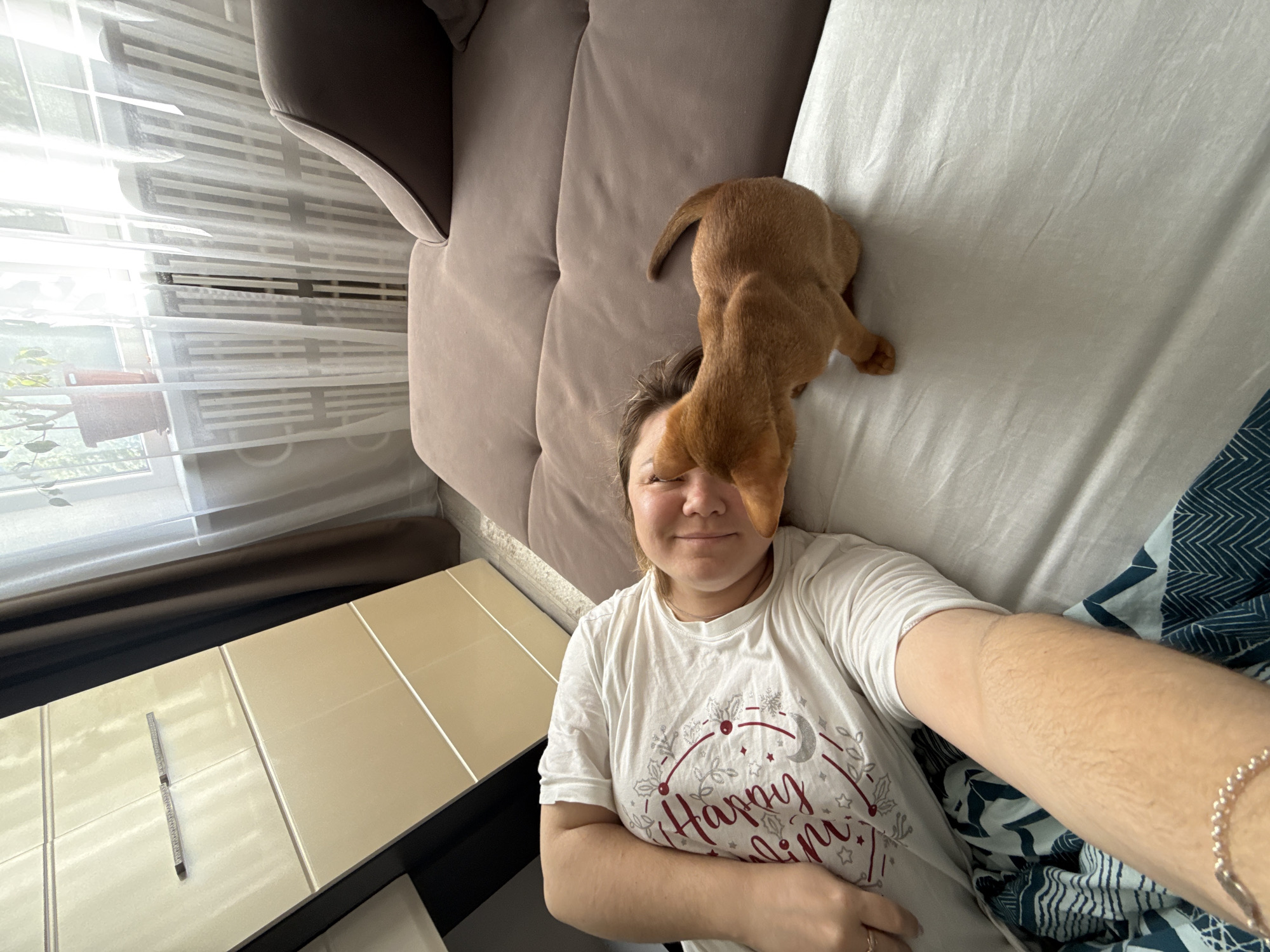 A woman is lying on a bed with a small, ginger dog nuzzling her face. She is wearing a white t-shirt with a festive design. The room appears to be a bedroom, with a sofa and a dresser visible. The lighting suggests it is daytime, possibly morning or afternoon. The background shows a window with sheer curtains and a radiator. The overall impression is one of domestic comfort and affection between the woman and her pet. No specific landmarks of Stary Oskol, Russia, are identifiable in this image.