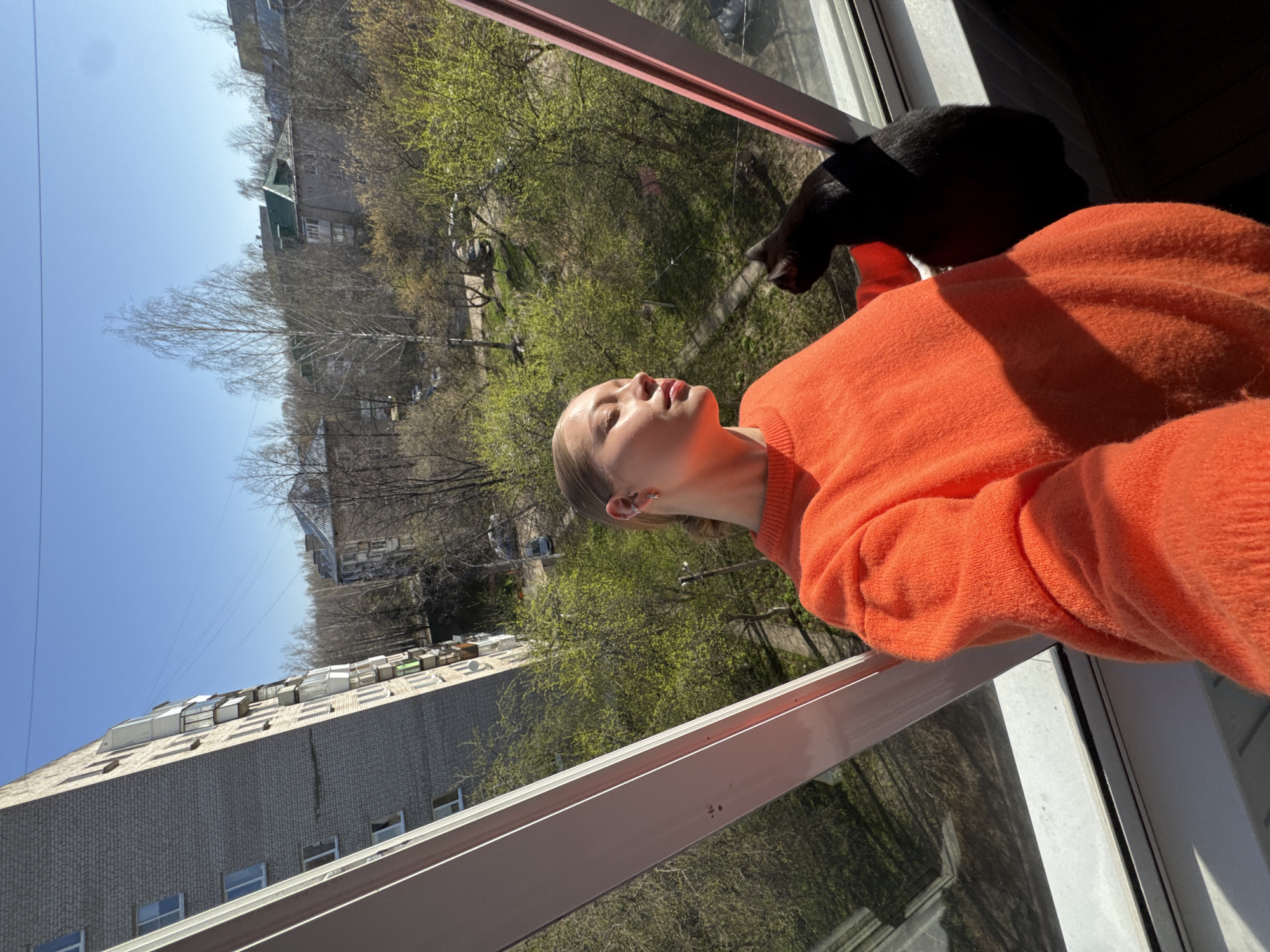 A vertical, eye-level shot shows a woman in an orange sweater standing by an open window with a black cat. The woman faces slightly upwards, with her mouth open. The window frame is light pink. Outside the window, green trees are visible and a brick building is seen in the distance under a clear blue sky. Based on the urban architecture, this location appears to be in the city of Chaikovsky, Russia.