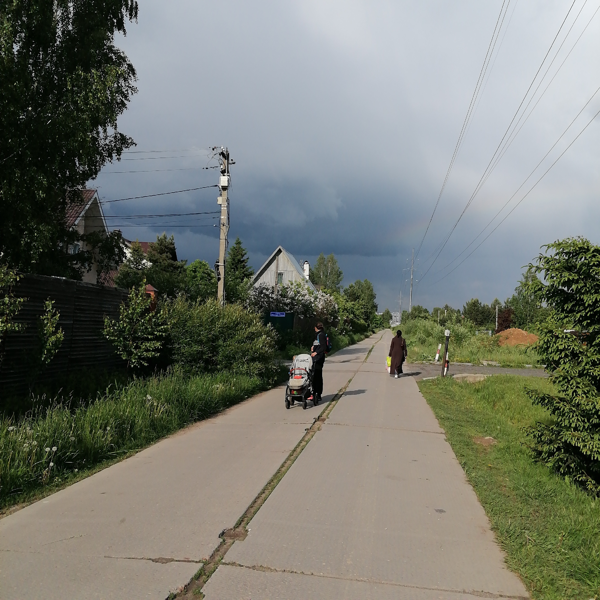 The image shows a paved road in Здравница, Russia. A person pushes a stroller along the left side of the road, while another person walks ahead on the right side. The road has a central groove with vegetation growing in it. Buildings and trees line the left side of the road, while grass and bushes are on the right. Overlapping powerlines are visible above. The sky is overcast with a patch of darker clouds in the distance. A faint rainbow is visible near the top center of the image.