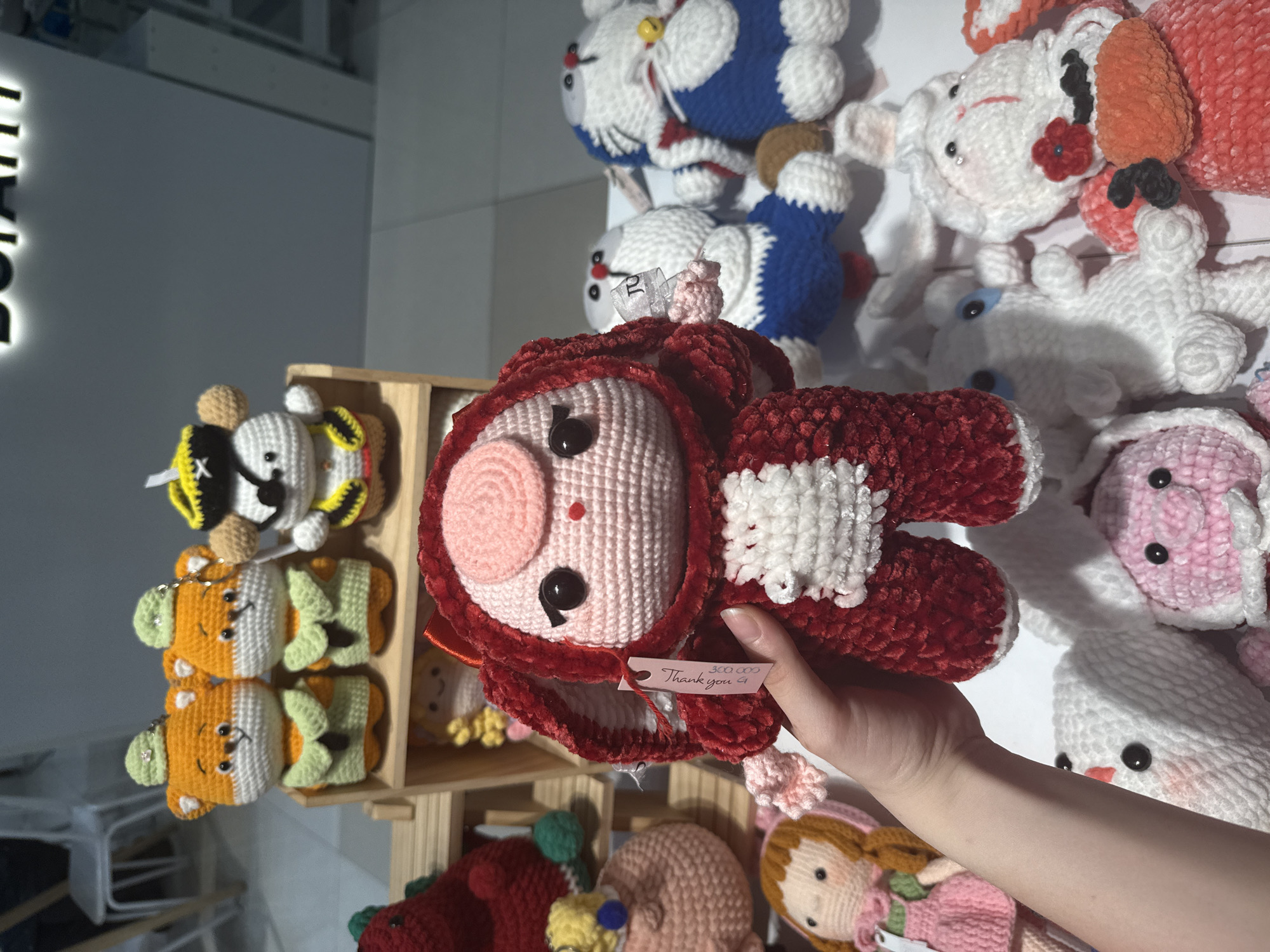 The image is an eye-level, interior shot featuring handcrafted crochet dolls displayed for sale. A hand holds up a reddish-brown crochet doll with a light pink face and a tag that says "Thank you" and indicates a price. Behind this, a wooden display case holds a variety of crochet animals including yellow and black bees, and orange fish. To the right, a wall is lined with more crochet dolls in shades of white, blue, and pink. The scene is brightly lit. In the background, blurred chairs and tables suggest a shop setting. The presence of Vietnamese text on the price tag suggests the location is Nha Trang ward, Vietnam.