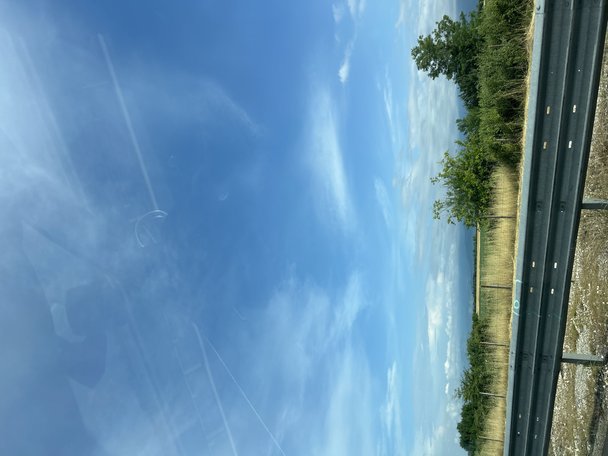 The image, captured from the interior of a vehicle, displays a bright, sunny roadside landscape, likely in the area of Malo Konare, Bulgaria. A grey metal crash barrier runs along the lower right side of the frame, indicating the edge of a road. Beyond the barrier, a strip of dry, golden-brown grass leads to dense green trees and bushes. Further in the background, agricultural fields, possibly of a cereal crop, extend towards hazy, distant hills or mountains.
The upper two-thirds of the image are dominated by a clear blue sky, adorned with scattered white clouds, ranging from wispy cirrus streaks to more defined cumulus formations. Reflections on the vehicle's window are noticeable on the left side, showing subtle outlines of the car's interior or perhaps a person, along with faint linear reflections that could be overhead power lines or contrails.
The scene depicts a daytime view under fair weather conditions, suggesting travel through a rural setting. On the crash barrier, a light blue spray-painted marking clearly shows the number "10", or possibly "L0", suggesting some form of road survey or maintenance identification.