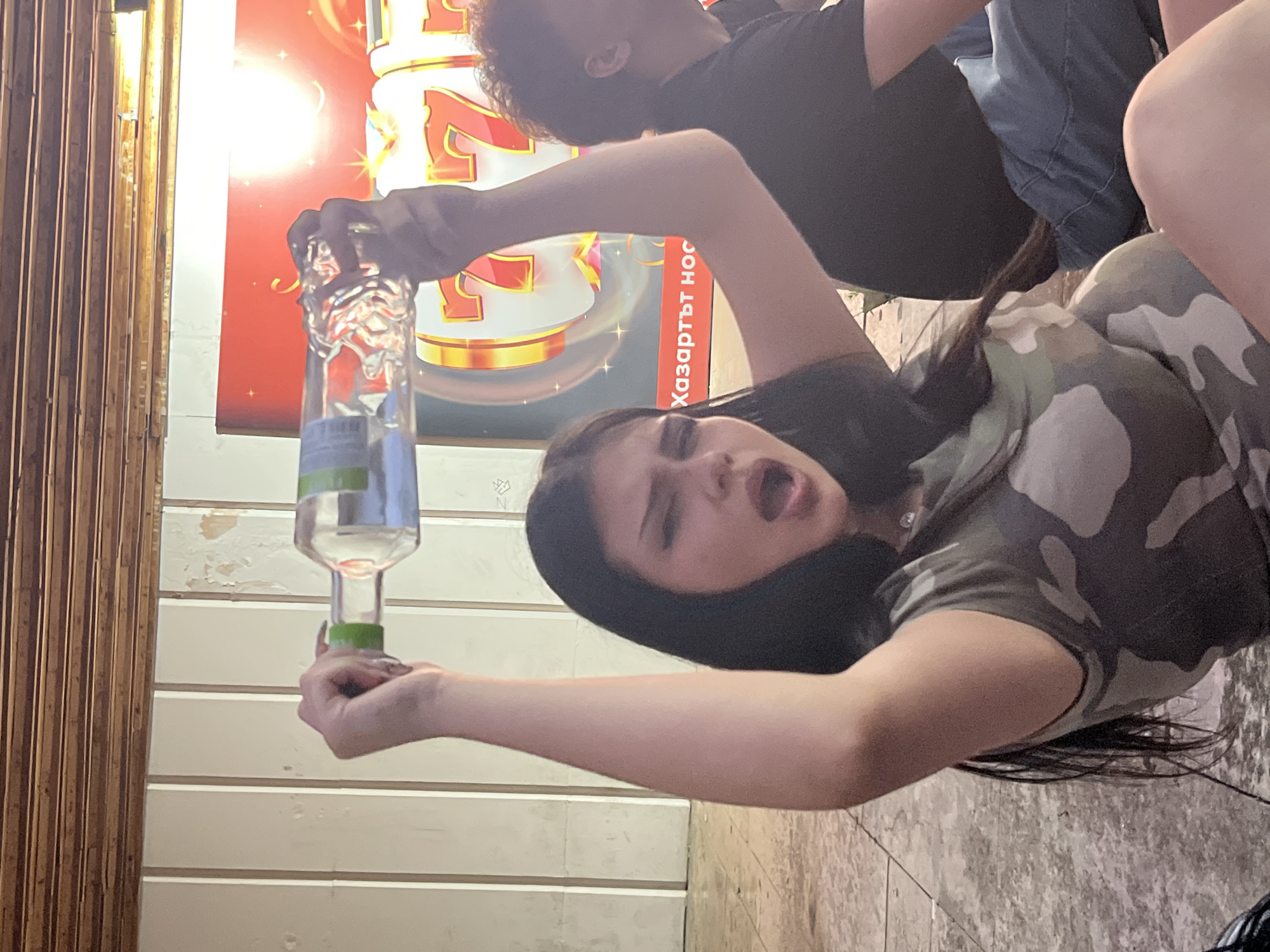 In Vratsa, Bulgaria, at street level, a young woman with dark hair, wearing a camouflage t-shirt, sits on the ground, holding a clear glass bottle filled with a clear liquid over her head. Her mouth is open, and her eyes are wide, suggesting an expression of surprise or excitement. To her right, a person wearing a black t-shirt and blue jeans is partially visible, also seated on the ground. Behind them, a large, brightly lit sign displays the numbers "777" in gold with red accents, accompanied by Bulgarian text that reads "Хазартът носи" (Gambling brings). The environment appears to be an outdoor public space with a tiled floor and a wooden ceiling overhead.