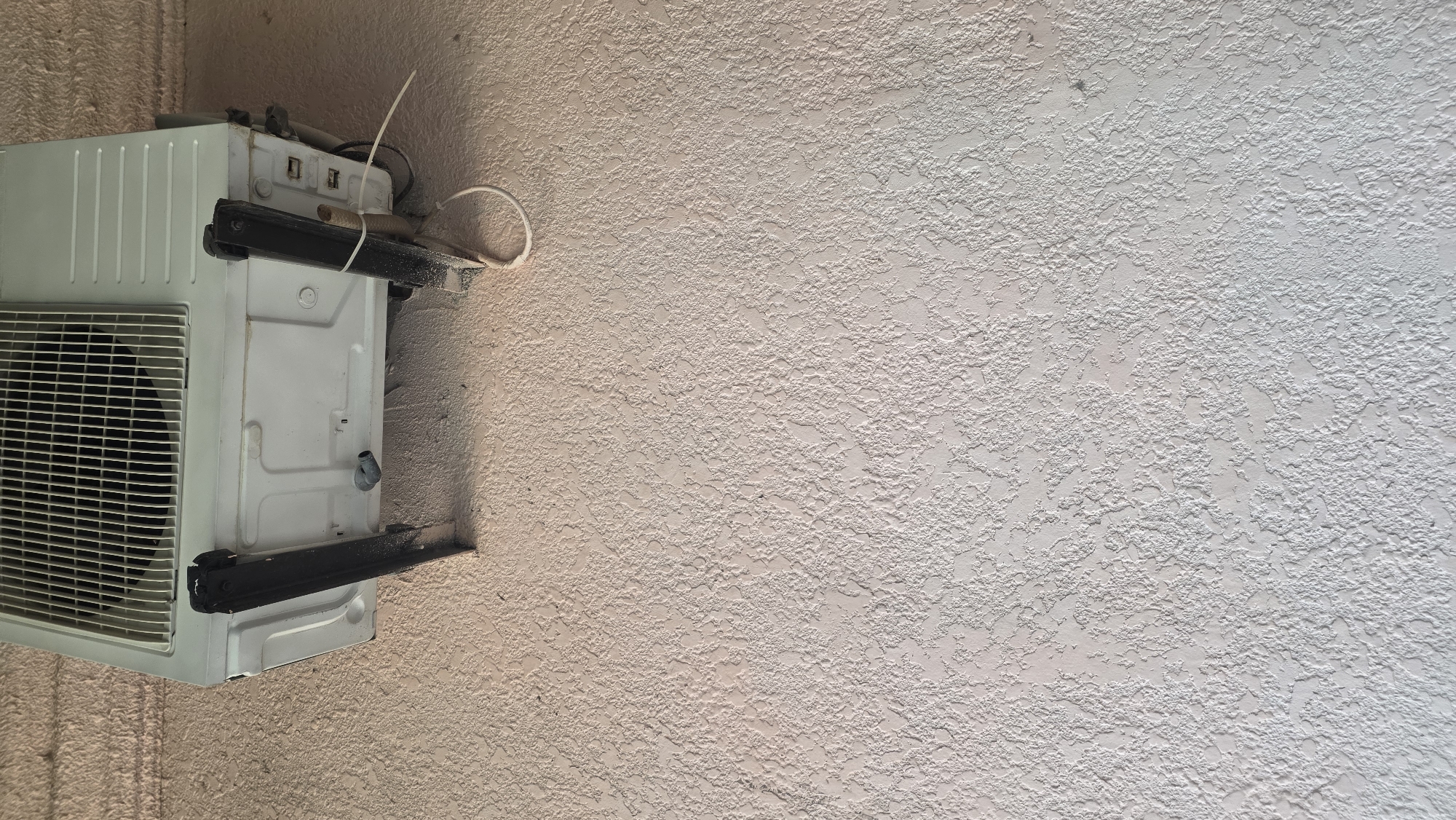 An outdoor shot shows a white air conditioning unit mounted on a textured, light pink wall. The unit is supported by two dark, angled brackets. A white cable hangs loosely from the unit. The scene is likely from an urban or suburban exterior in Chuy, Kyrgyzstan.
