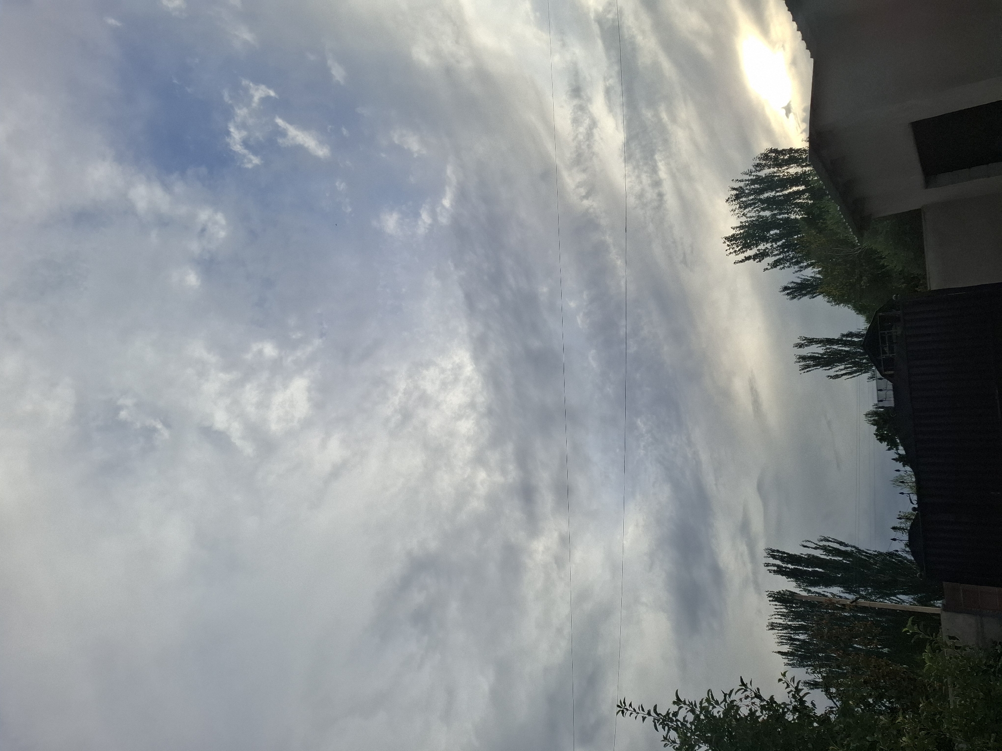 The image shows a sky with scattered clouds, some of which are thin and wispy, while others are thicker and more opaque. The sky is a mix of grays and blues, with a patch of brighter light on the right side, suggesting the sun is partially obscured by clouds. In the foreground, there are trees with dark green foliage, some tall and slender, and others with a fuller canopy. A dark fence runs across the middle ground. On the right side of the image, a portion of a light-colored building with a visible doorway is present. The scene appears to be an outdoor setting, possibly a residential area or a yard. There are no people visible in the image. The environment suggests an overcast or partly cloudy day, with the sun attempting to break through. The presence of trees and a building indicates a developed area, consistent with a location within a city or town.