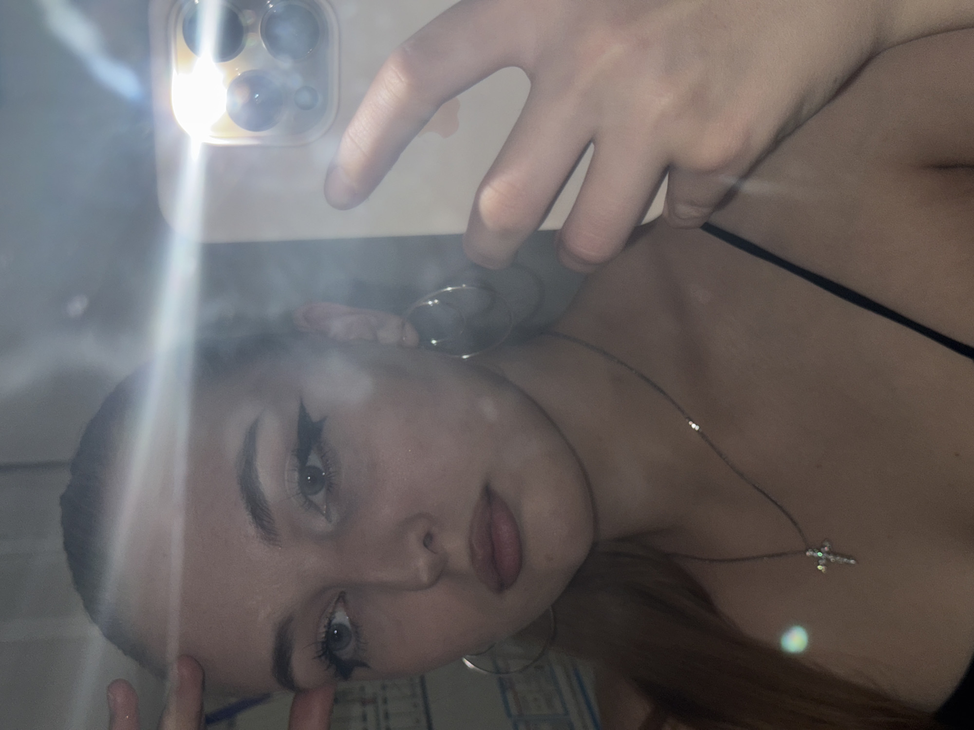 A close-up, selfie-style image shows a light-skinned person with dramatic eyeliner. The person has light-colored eyes, and a cross necklace. They are holding a phone up to take the picture, the flash is shining brightly in the upper left of the image. The image is tilted counterclockwise. No location information or actions are depicted. There are no signs of the city of Perm, Russia.