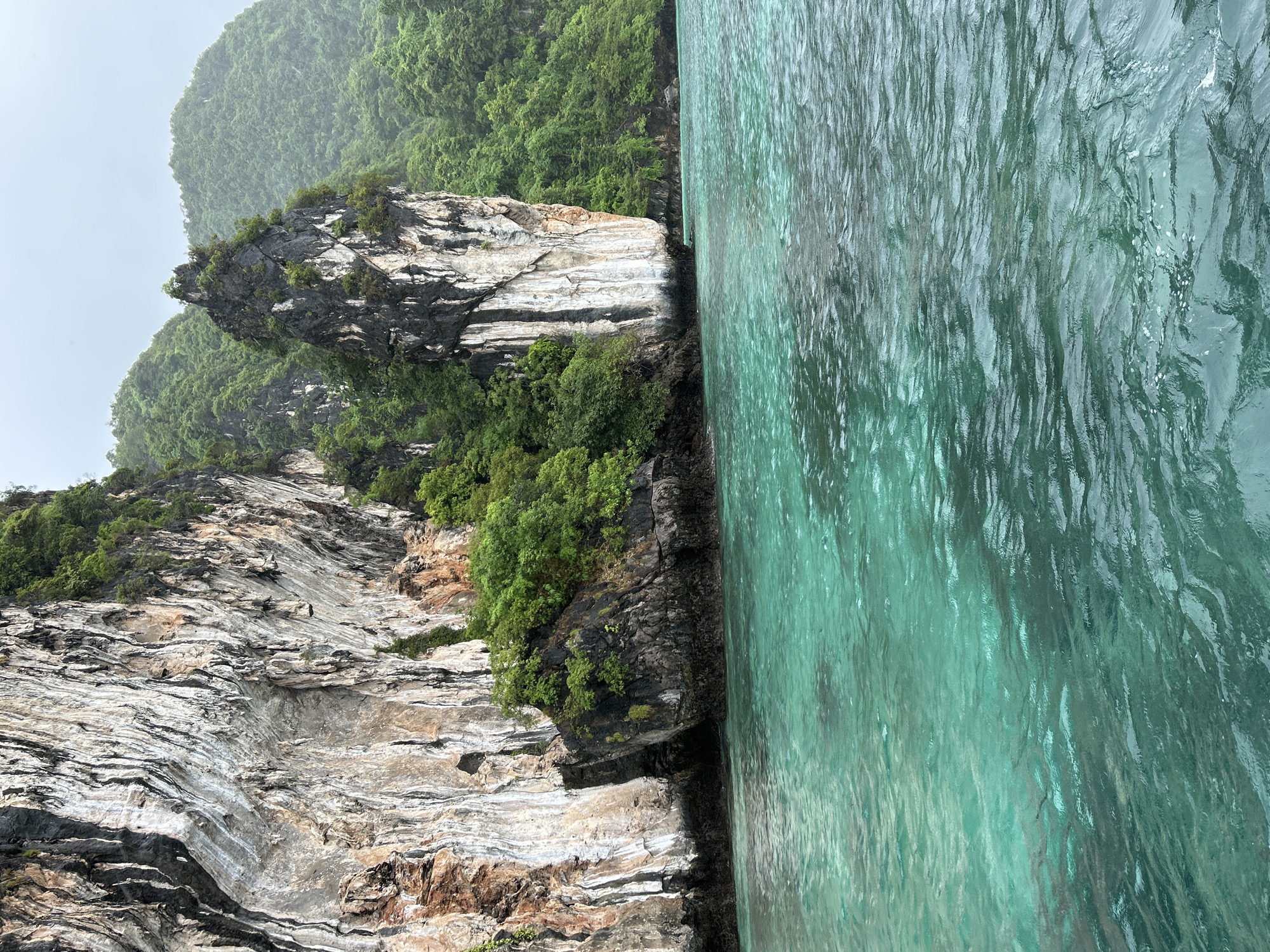 The image shows a view of the ocean near Haad Hin Khom, Thailand. A large, sheer limestone cliff face with visible strata and patches of green vegetation dominates the left side of the frame. To the right of this cliff, a smaller, darker, and more jagged rock formation juts out from the water, also covered in dense green foliage. The background is filled with lush, rolling hills covered in trees, suggesting a tropical environment. The water in the foreground is a clear, vibrant turquoise with gentle ripples, indicating calm seas. The sky is overcast and a pale grey, hinting at a cloudy or potentially rainy day. No people or man-made structures are visible in the scene, focusing entirely on the natural landscape.