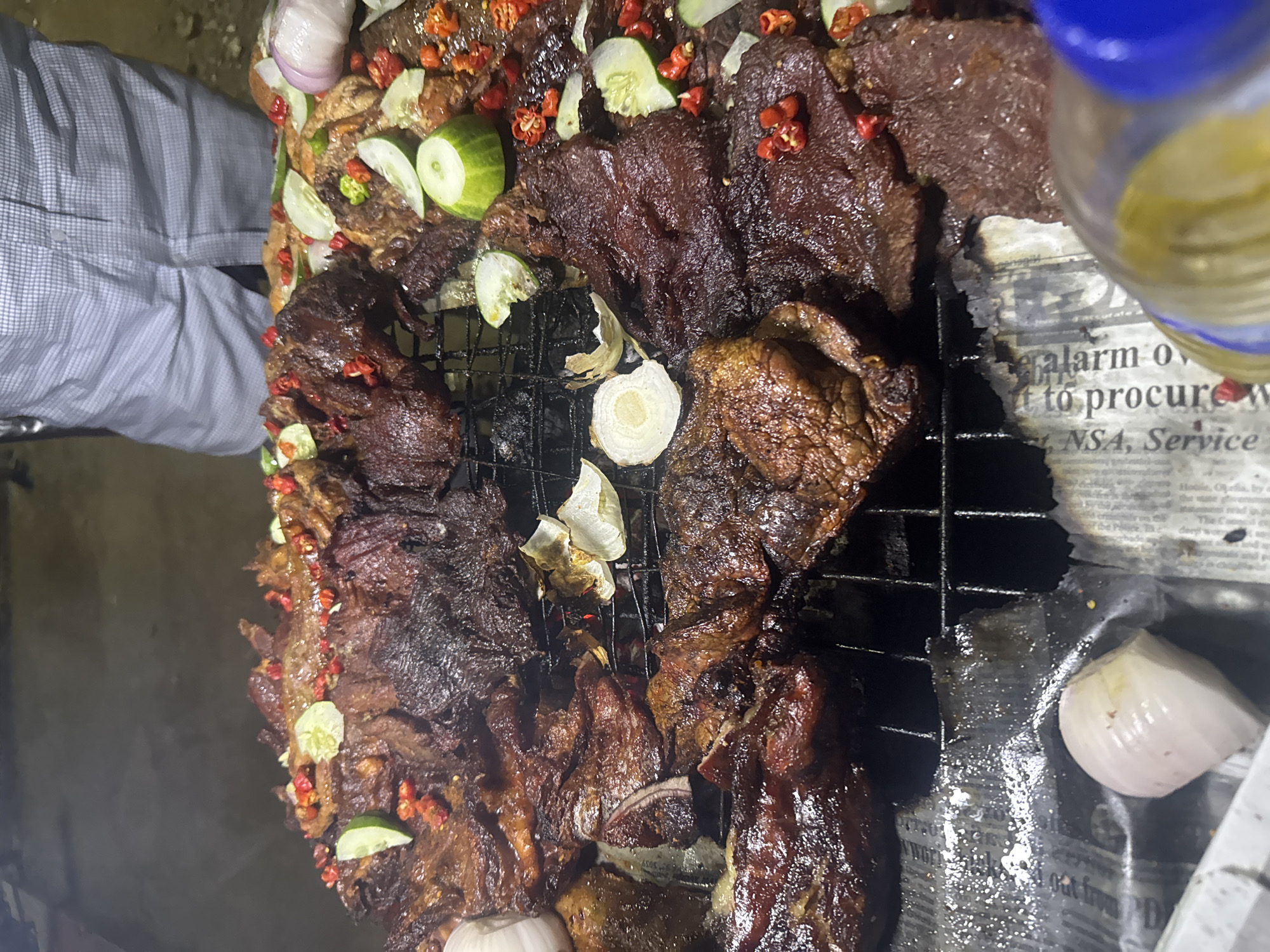 The image shows a close-up, overhead view of grilled meat being prepared or served, likely in Abuja, Nigeria. The meat, a dark, rich brown color, is arranged on a metal grill. It appears to be beef, cut into various sizes and shapes, with some pieces having a slightly charred texture. Scattered amongst the meat are slices of cucumber, red chili peppers, and what seem to be pieces of onion. The grill itself is placed over what looks like a bed of newspaper, which is stained with grease from the cooking process. To the left of the frame, a person's legs are visible, wearing a light blue and white checkered shirt. In the upper left corner, parts of an onion and a few chili peppers are also visible. In the upper right corner, a blurry glass bottle with a blue cap and a yellow liquid is partially visible. The overall lighting suggests it might be evening or indoors with artificial light. The presence of newspaper as a base and the rustic arrangement of the food might indicate a street food setting or a casual outdoor preparation. There is some visible text on the newspaper, which appears to be a news article, but it is largely obscured by grease and the angle. Some legible words include "alarm o", "to procure w", "NSA, Service".