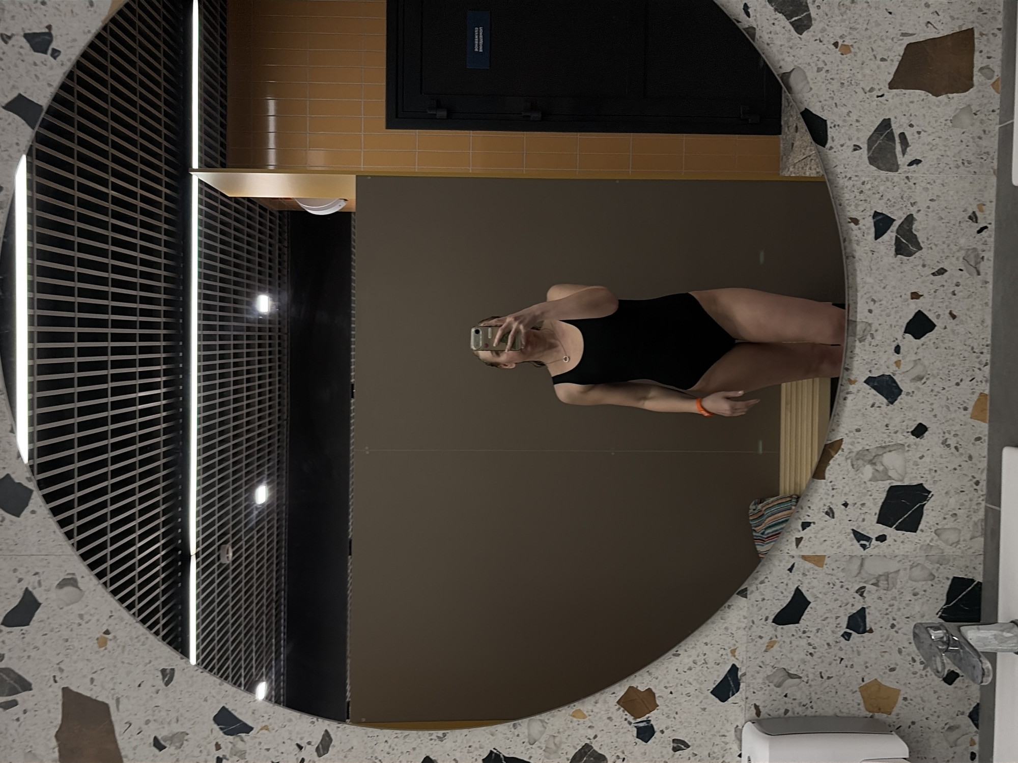 The image captures a mirror selfie taken by a female subject in what appears to be a changing room or restroom of a public facility, likely a swimming pool or gym, located in Votkinsk, Russia.
**Scene Description:**
The central subject is a female, seen from the chest to the upper thighs, reflected in a large, circular mirror. She is wearing a plain black one-piece swimsuit. Her face is partially obscured by the light-colored smartphone she holds with both hands to take the photograph. She wears a simple silver-toned necklace and an orange wristband on her right wrist.
**Environment:**
The mirror features a distinctive speckled frame composed of white, grey, black, and ochre-colored fragments, resembling terrazzo. In the immediate foreground of the reflection, a white sink with a chrome faucet and a white wall-mounted soap dispenser are visible on the left. The background reflected in the mirror reveals a neutral grey-brown partition or wall directly behind the subject. To the right, a section of wall is covered in orange-brown square tiles. A dark-colored door, possibly black or dark grey, is set within the orange-tiled wall. A white sign with white Russian text is affixed to this door, which reads "ПОМЕЩЕНИЕ СЛУЖЕБНОЕ" (SERVICE ROOM / STAFF ONLY), indicating restricted access. Above the partitions, a modern grid-patterned ceiling with multiple linear fluorescent lights and recessed spotlights is reflected. A multi-colored striped item, possibly a towel or clothing, rests on a low bench or ledge in the reflection, to the left of the subject. A white, wall-mounted dispenser, likely a paper towel dispenser or hand dryer, is partially visible on the orange-tiled wall.