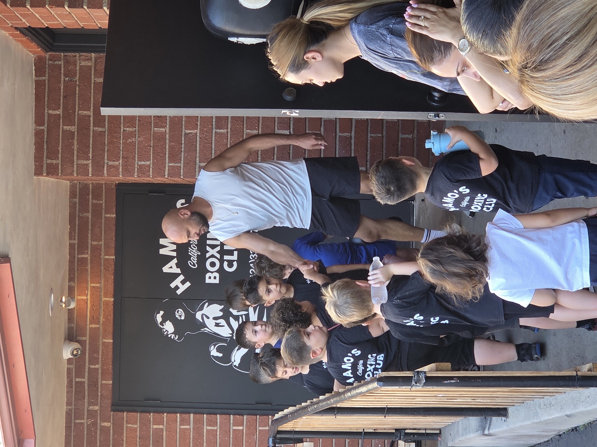 A group of young boys and an adult male are gathered outside a building with "HAMO'S CALIFORNIA BOXING CLUB" written on the wall. The adult male, wearing a white sleeveless shirt and black shorts, is standing and looking down at one of the boys. Many of the boys are wearing black t-shirts with the boxing club's logo. Some of the boys are holding water bottles. A young girl with brown hair is also visible in the foreground, facing away from the camera. The building appears to be made of brick, and there are two security cameras mounted on the wall above the entrance. The image was taken on a sunny day.