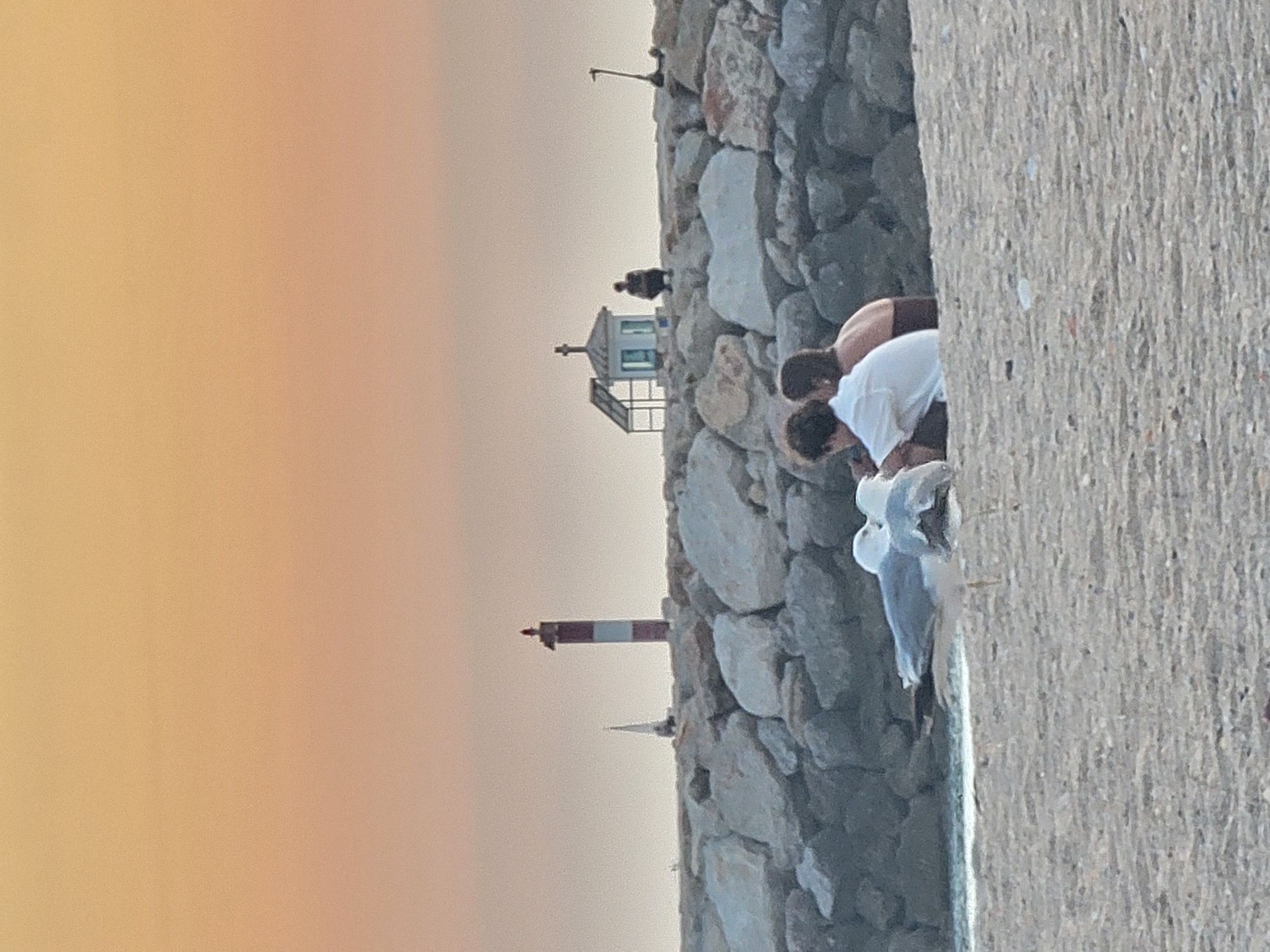 In Quarteira, Portugal, two individuals are seated on a breakwater, facing a small, white structure with a cross on top, possibly a memorial or shrine. They are positioned in front of a rocky embankment, with a lighthouse and a sailboat visible in the distance. Two seagulls are in the foreground on the sandy beach, appearing to interact. The sky exhibits a gradient of orange and yellow hues, indicating either sunrise or sunset. An electric scooter is also present on the breakwater.