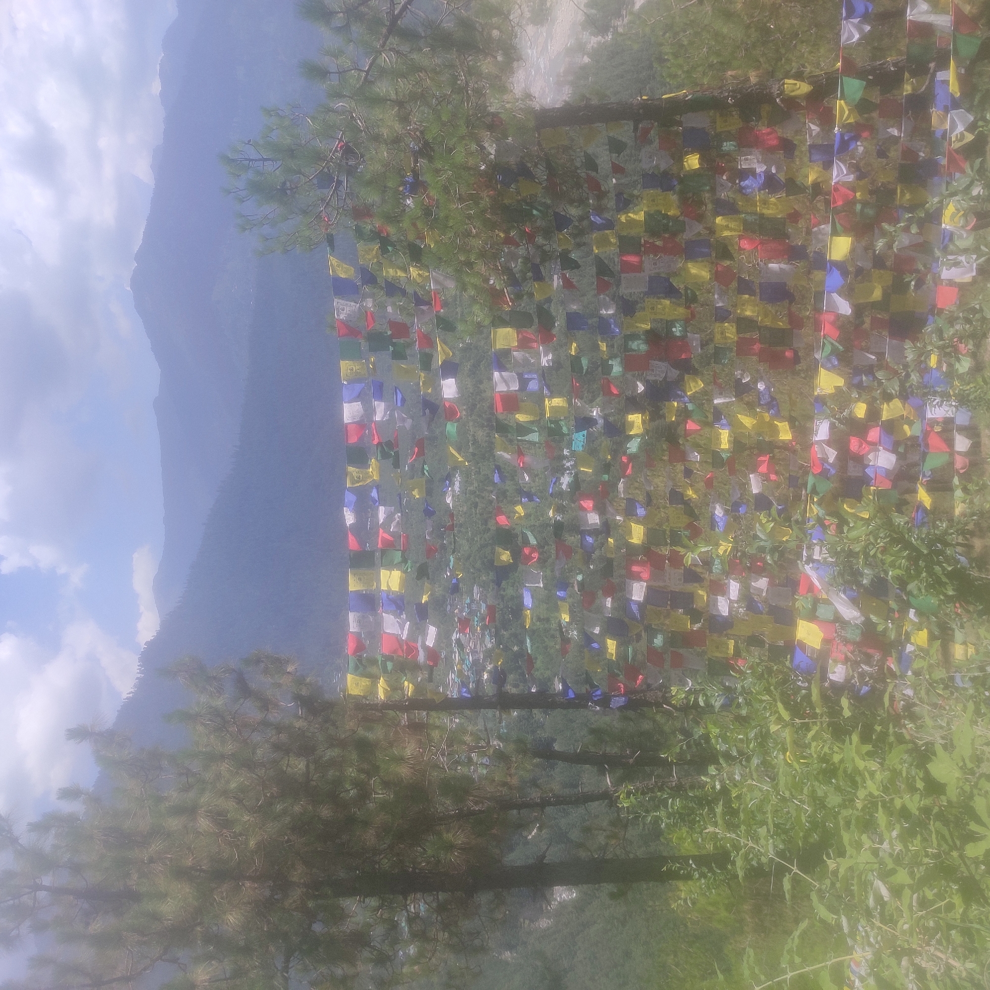 This is a photograph taken from a high vantage point overlooking the town of Baragran, India. In the foreground, a string of colorful prayer flags, a common sight in Buddhist regions, is draped across the frame, obscuring a portion of the view. Beyond the flags, the town of Baragran is visible nestled within a lush, green valley. Buildings with red and grey roofs are spread across the landscape. In the background, rolling mountains rise, fading into a hazy, blue-grey sky with scattered clouds. The overall impression is one of a serene, mountainous landscape with a touch of cultural vibrancy from the prayer flags.