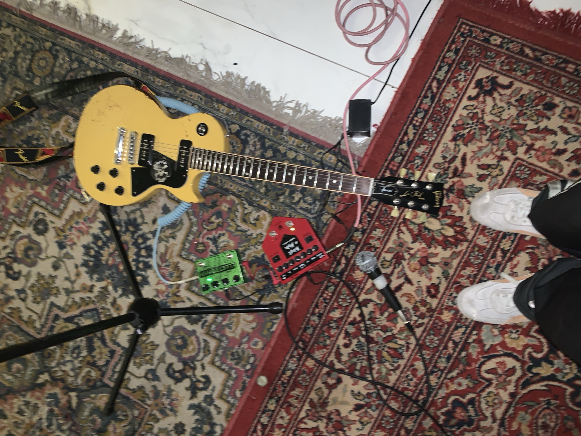 The image depicts a musical setup indoors. A yellow electric guitar rests on a stand. The guitar has a black pickguard with a skull design. There are two guitar effects pedals, one green and one red, positioned near the guitar stand. A microphone is also visible, lying on a patterned rug. A pair of white sneakers with black pants are on the right. The location is unknown; there is nothing in the image to identify it.