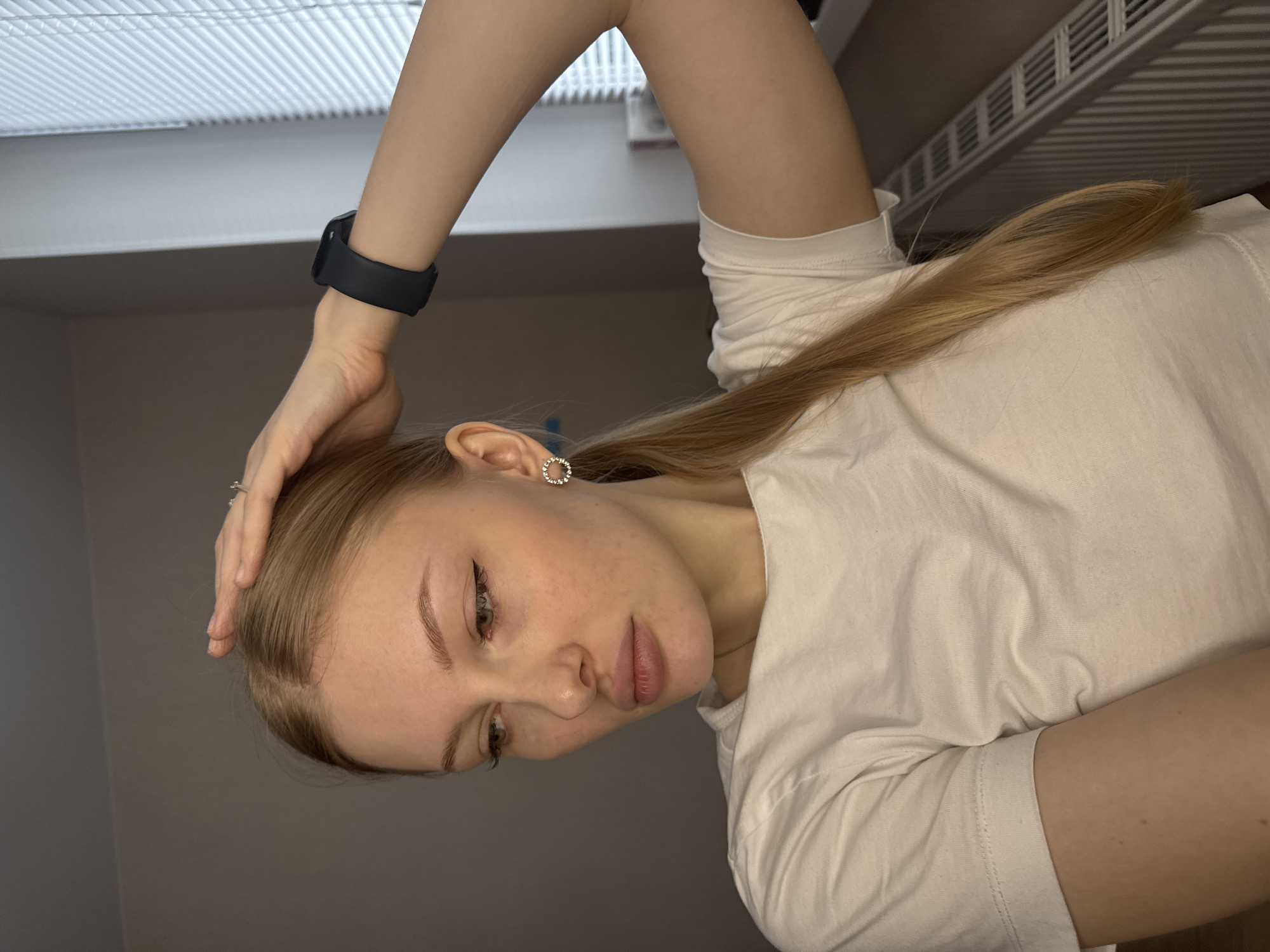 The image shows a young woman, likely in her late teens or early twenties, captured indoors in Perm, Russia. She has long, straight, light brown hair, parted to one side, and fair skin, with visible eyebrows and some subtle makeup, including eyeliner. She is wearing a white t-shirt.
On her left wrist, she wears a black smartwatch or fitness tracker. A small, circular, silver-colored earring, adorned with tiny clear stones, is visible on her right ear, and she has a ring on her left ring finger. Her left hand is raised to her head, gently touching her hair. She gazes downwards and to her right, displaying a neutral to slightly contemplative expression.
The background consists of a light-colored, possibly white, surface with horizontal ribbed patterns in the upper part of the frame, which might be a ceiling vent or a light panel. The lower portion of the background is a plain, dark gray wall. The lighting appears to be artificial and even, casting a soft glow. No visible text is present in the image.