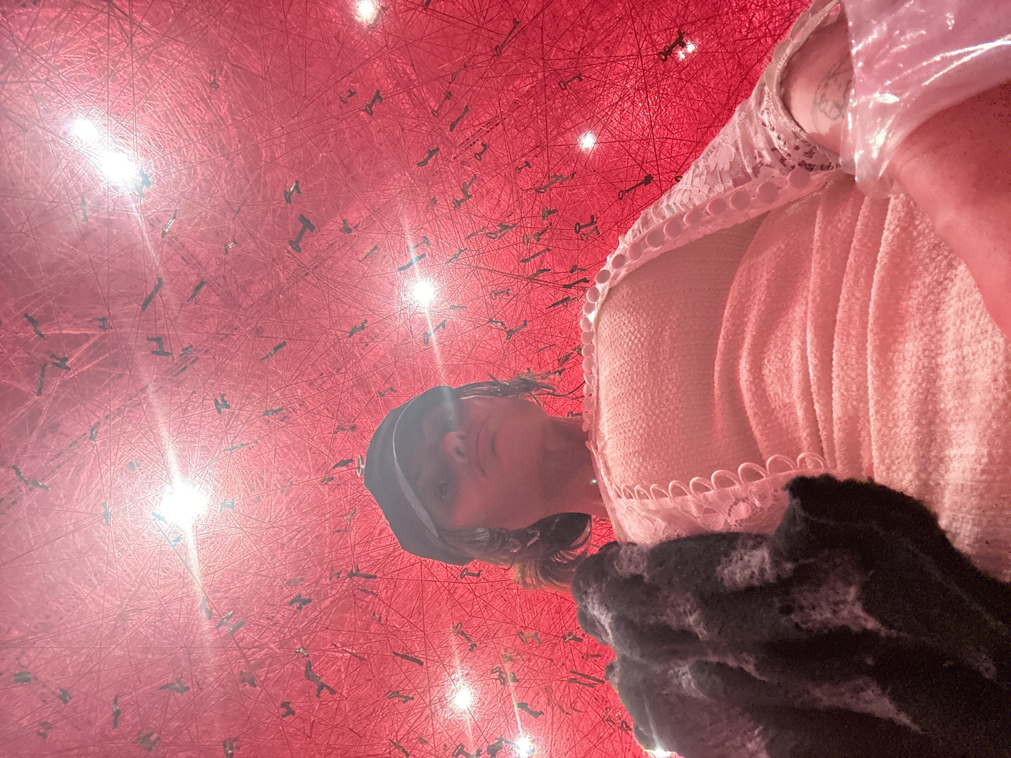 The image captures a unique perspective of a person interacting with a vibrant art installation. The photograph is taken from a low angle and is rotated, presenting the subject on the right side of the frame, partially visible from the chest up. The person has dark hair and is wearing a dark hat or headband. They gaze directly at the camera with a neutral to contemplative expression. Their attire includes a light-colored, possibly pale pink or off-white, top with a textured fabric and a decorative lace or ruffled trim along the neckline and shoulder, featuring small buttons. A dark, possibly knitted or furry, garment or accessory is visible in the bottom right corner.
The background is dominated by an immersive art installation, characterized by an intricate, dense web of crisscrossing red threads that fill the entire space. Numerous small, dark metallic keys are suspended at various points within this elaborate red network, creating an intriguing visual texture. Several bright, star-like lights are embedded within or reflected by the threads, adding points of illumination to the scene. The overall atmosphere suggests an indoor art exhibit or gallery in London, United Kingdom, where a visitor is engaging with the immersive environment. There is no visible text within the image.