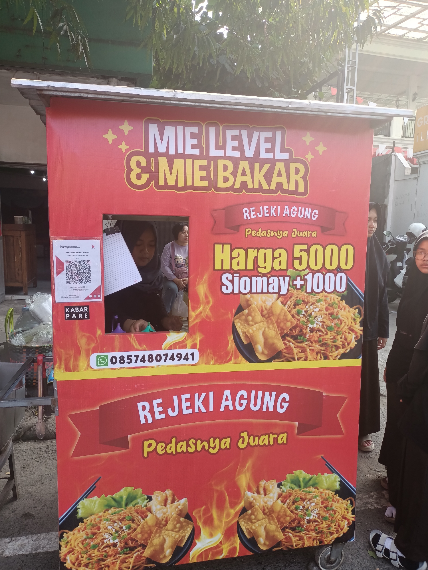 A red food cart branded with "MIE LEVEL & MIE BAKAR" is set up outdoors in what appears to be a street setting in Pare, Indonesia. The cart features prominent text advertising "REJEKI AGUNG" and the slogan "Pedasnya Juara" (Spiciness is Champion). Below this, a price is displayed: "Harga 5000 Siomay +1000" (Price 5000 Siomay +1000), accompanied by an image of a noodle dish with dumplings. A QR code for payment is affixed to the left side of the cart, along with a WhatsApp number, "085748074941". Inside the cart, two individuals, likely employees, are visible behind a small window, with one serving a customer. The surrounding environment shows a building in the background and some vegetation, suggesting an urban or commercial area. The cart is mobile, equipped with wheels.