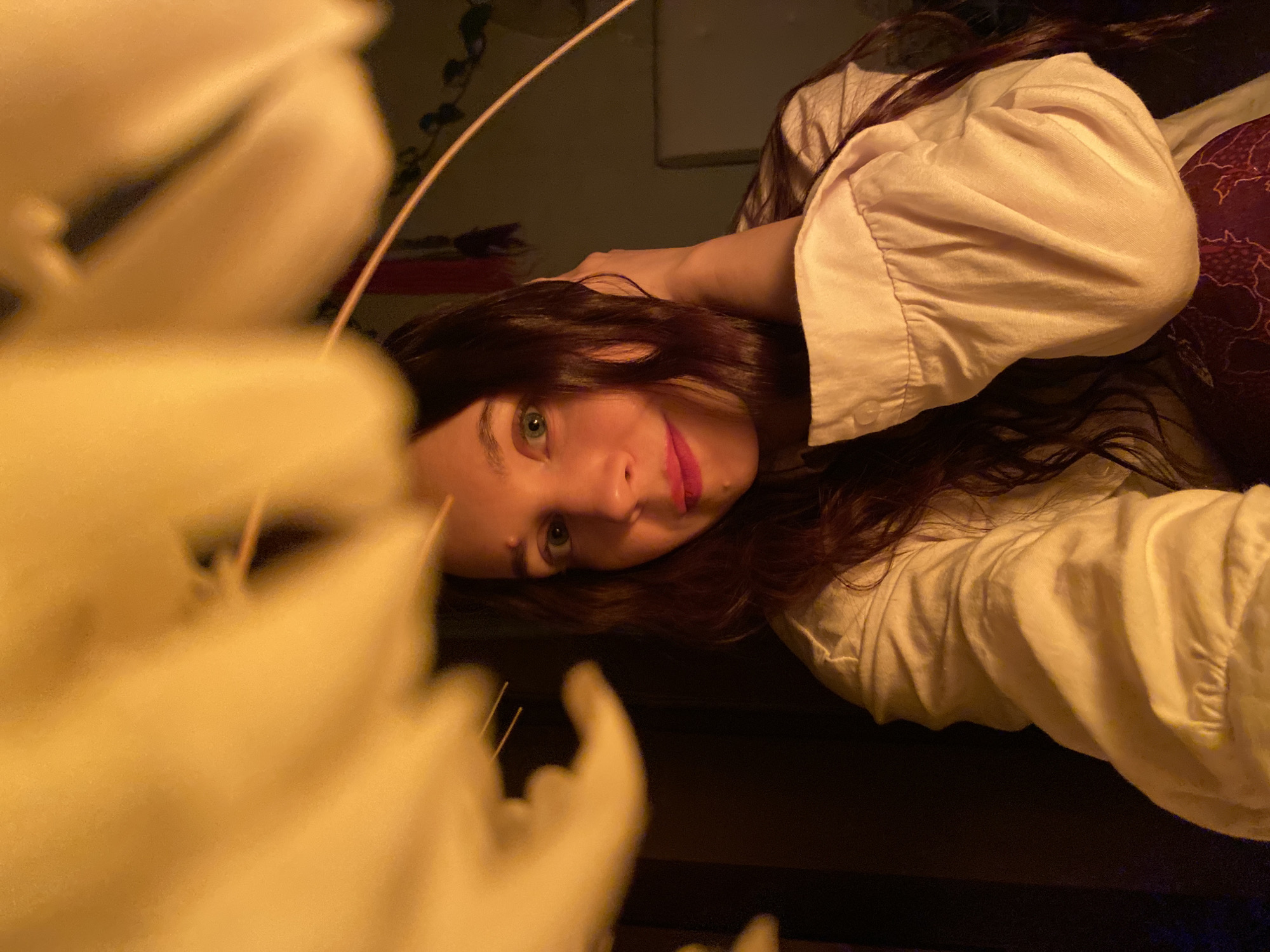 A person is lying down, facing the camera. She has long brown hair, blue eyes, and pink lipstick. She is wearing a white shirt with rolled-up sleeves. Her right hand is behind her head. A blurry, off-white object, possibly a flower or decorative item, is in the foreground, partially obscuring the view. The background appears dark and somewhat indistinct, with a possible plant visible in the upper portion. There is no indication that the photo was taken in Chaikovsky, Russia.