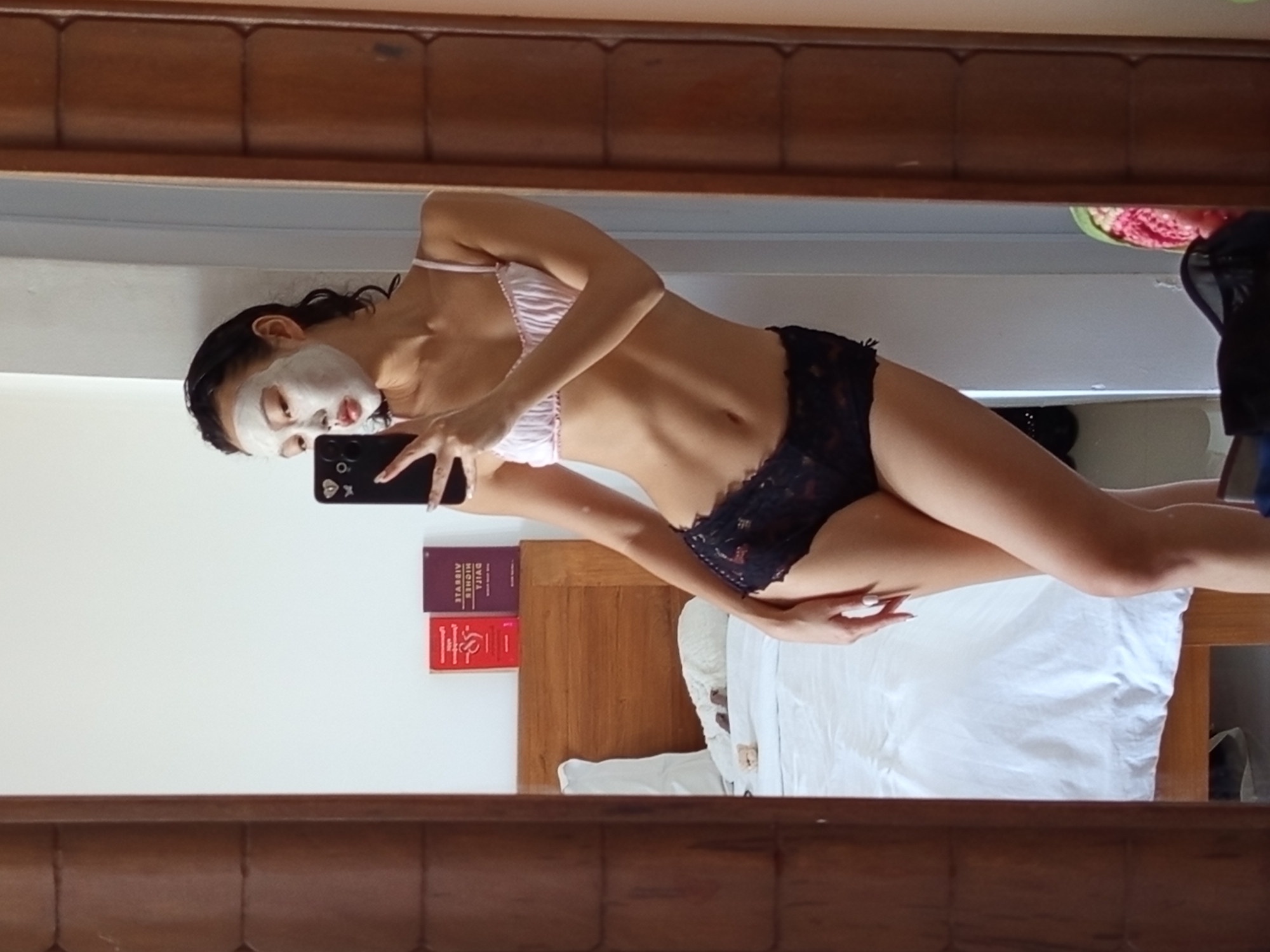 A mirror image depicts a person in lingerie with a face mask, holding a smartphone, seemingly taking a selfie. The backdrop suggests a bedroom setting with a bed and possibly books. There is no indication of the location of North Kuta, Indonesia.