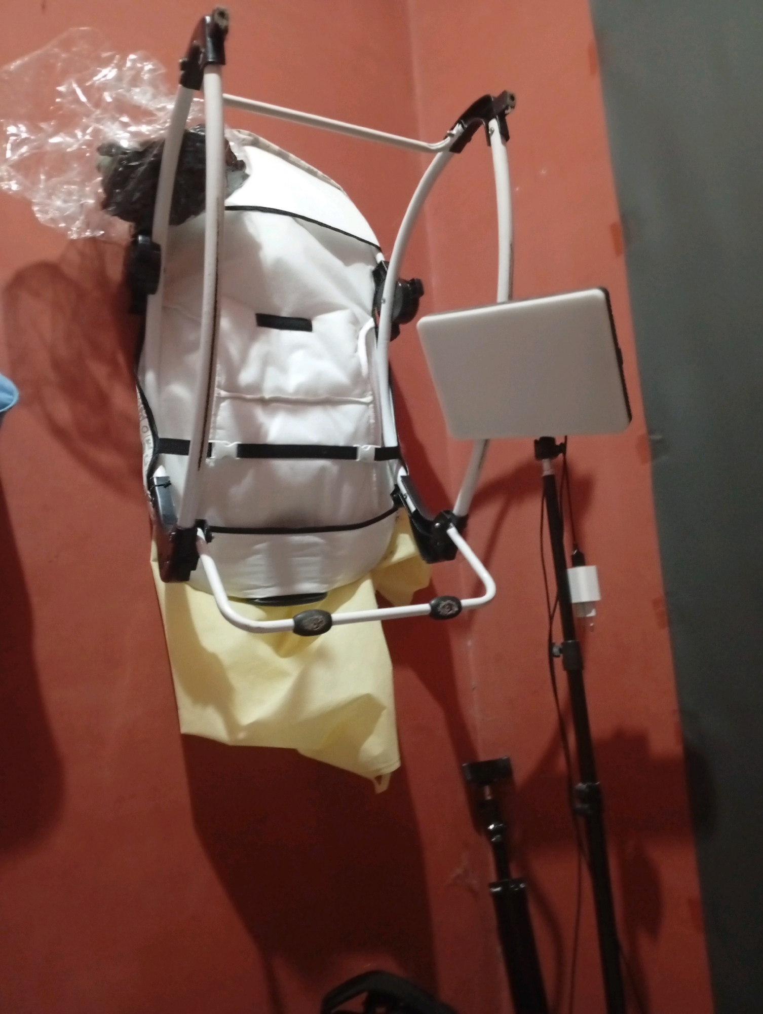 The image depicts an indoor scene, likely a room in Tasikmalaya, Indonesia. The dominant color of the wall is a reddish-brown, with a darker grey section on the right. In the center of the image, a white baby swing or bassinet is hanging against the wall. It has black accents and a pale yellow fabric underneath. To the right of the swing, a black stand is visible, supporting a flat, white object, possibly a tablet or a small screen, attached at an upward angle. Wires and a white adapter are connected to the stand, suggesting an electronic device is in use or set up. The overall lighting is dim, casting shadows on the wall.