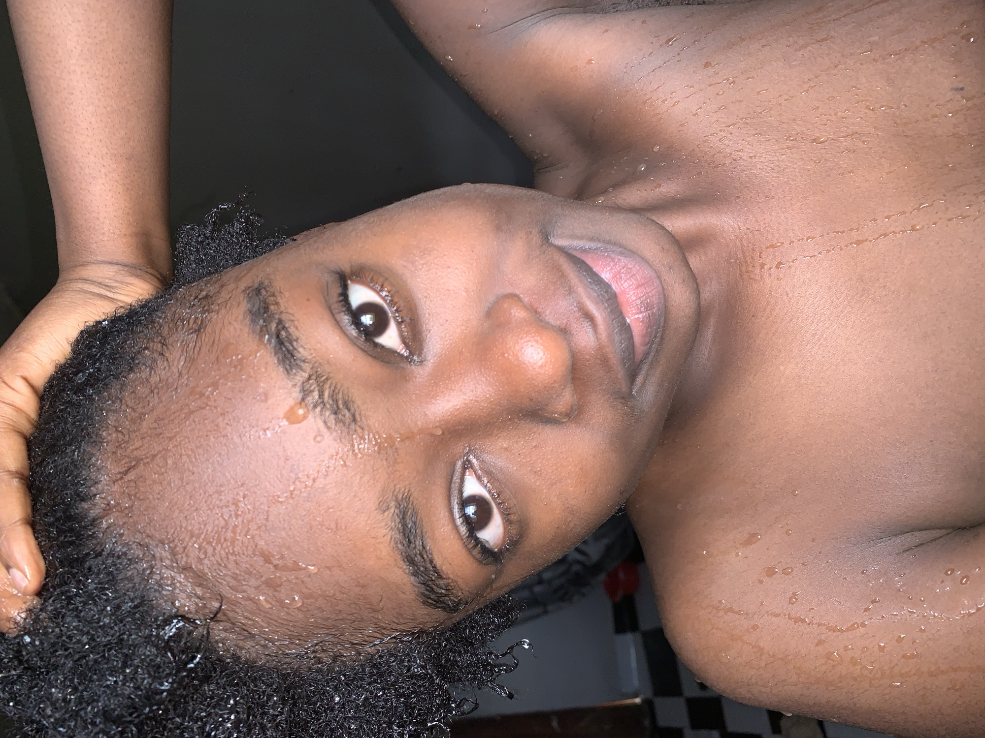 A young Black woman is shown from the chest up, with her head tilted back and her hand resting on her head, partially obscuring her hair. Her skin is wet, with visible water droplets on her forehead, temples, neck, and shoulders. She has dark, curly hair. Her eyes are open and looking towards the camera, and she has a slight smile. The background is dark, with a hint of a tiled wall visible on the left side. The overall impression is that of a person who has recently showered or been exposed to water. The image does not contain any information that would allow for its location to be determined to be in the city of Rafin Par, Nigeria.