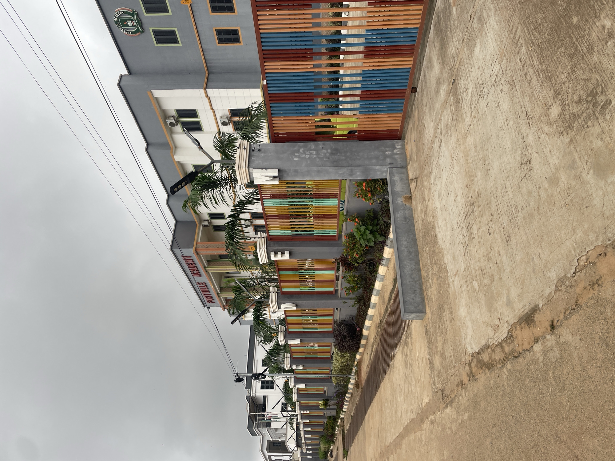 This image depicts the entrance to Fortville Academy, located in Abuja, Nigeria. The academy building, a modern structure with grey and white facades, is visible in the background. A prominent sign above the entrance reads "FORTVILLE ACADEMY" in red lettering. The academy is secured by a vibrantly colored fence, featuring vertical bars in shades of red, orange, blue, and yellow. The fence is supported by concrete pillars, some of which have the letters "F," "A," and "M" inscribed on them. Lush greenery and palm trees surround the entrance, enhancing the aesthetic appeal of the academy. The foreground shows a paved walkway and road, suggesting an urban setting. The sky is overcast, indicating a cloudy day. There are no people or vehicles visible in the image.