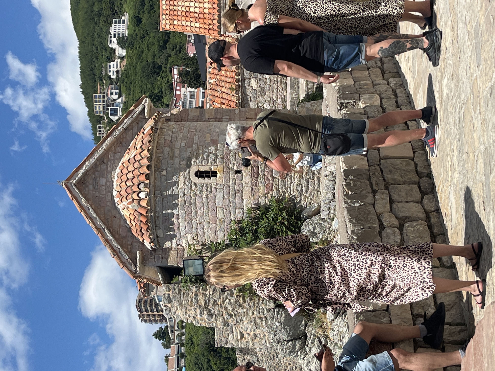 The image depicts a group of tourists standing on a stone-paved pathway in front of a stone building with a red-tiled roof. The building appears to be a historical structure, possibly a church or a fortress, with rough-hewn stone walls and a small arched window. Behind the building, a lush green hillside is visible, dotted with modern white buildings and apartment complexes. The sky is blue with scattered white clouds.
In the foreground, a woman with long blonde hair wearing a leopard-print dress is standing facing away from the camera, holding a pink object. To her left, a man in blue shorts and a dark shirt is seated on a stone wall, looking at his phone. To the right of the woman, a man with graying hair, wearing a green t-shirt and denim shorts, is taking a photograph with a vintage camera. A black bag is slung across his shoulder. Further to the right, a man in a black t-shirt and blue shorts, wearing a baseball cap, stands with his back to the camera. Beside him, a woman in a leopard-print off-the-shoulder dress is also looking away. The man in black has a large tattoo on his left leg.
The overall scene suggests a tourist area in Budva, Montenegro, likely within the Old Town or a similar historical site, given the architectural style of the building and the surrounding landscape. The presence of tourists engaged in photography and leisure activities indicates a daytime scene during warm weather.