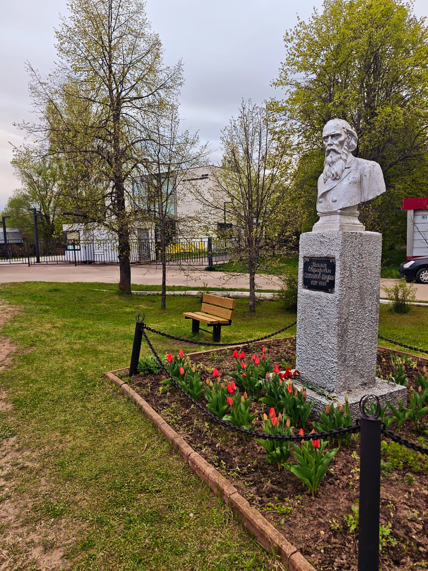 The image shows a bust of a man with a beard, placed on a stone pedestal. The pedestal has a plaque with Cyrillic text. Red tulips are planted around the base of the pedestal, enclosed by a low brick border and a chain fence. Behind the monument is a wooden bench, trees with budding leaves, and a building in the background. The setting is a grassy area, likely a park or memorial garden in Spas-Ugol, Russia.