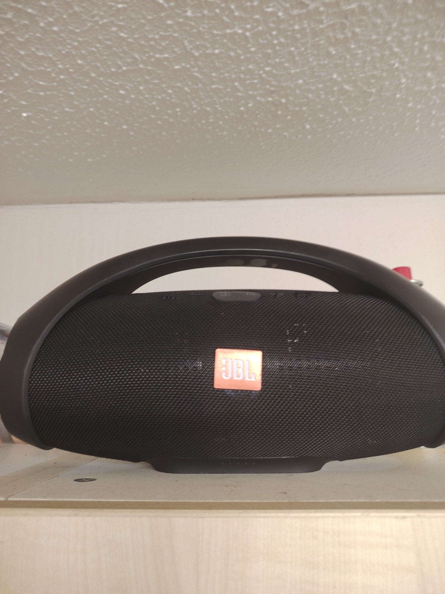 A black JBL Boombox portable Bluetooth speaker is centered on a light-colored wooden surface in an indoor setting in Huntingdonshire, United Kingdom. The speaker features an arch-shaped handle and a textured mesh grille with an orange JBL logo prominently displayed in its center. Behind the speaker is a plain light-colored wall. The upper portion of the image reveals a white ceiling with a distinct popcorn texture. No individuals or dynamic actions are present in the scene.