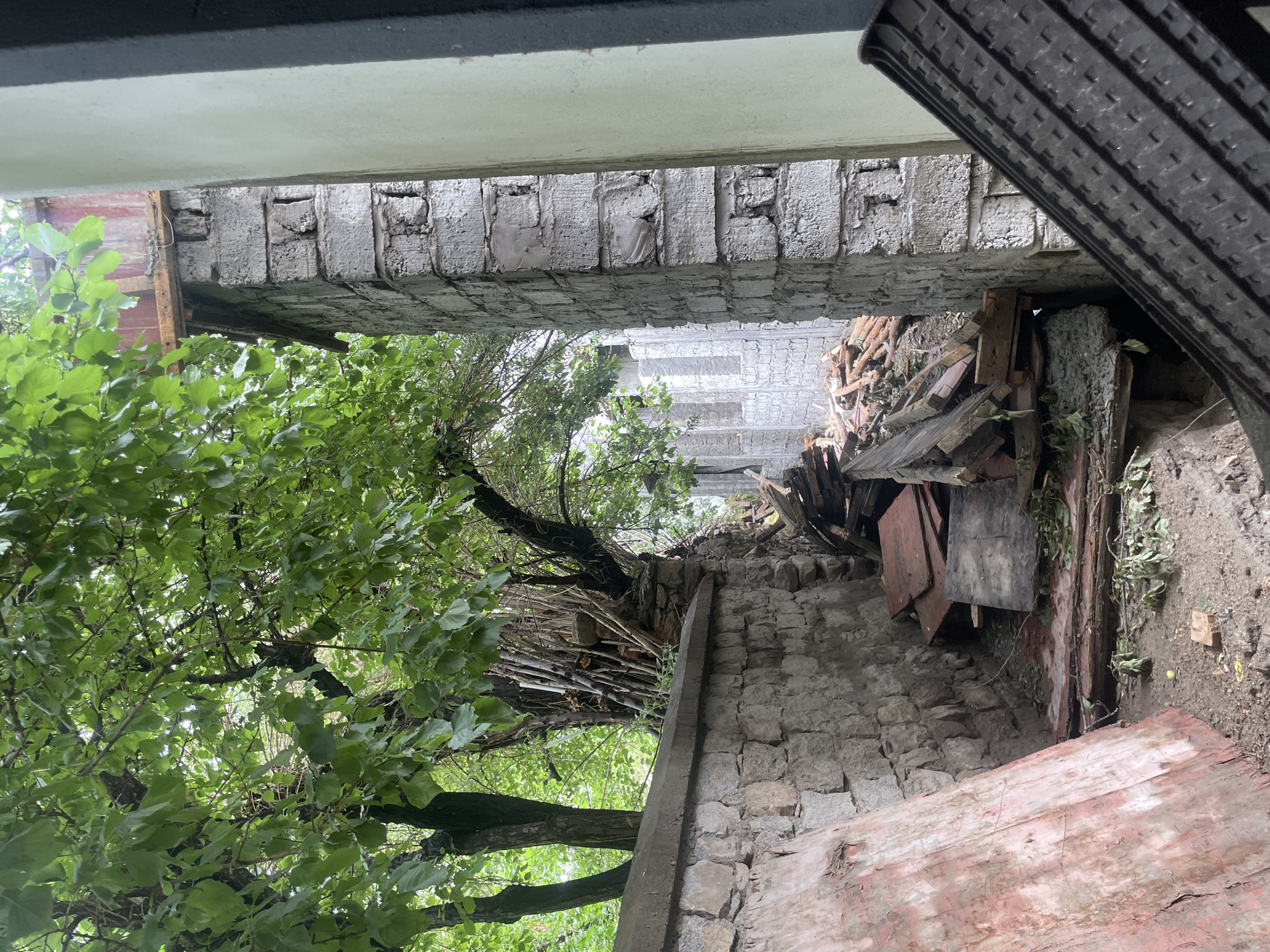 An outdoor, eye-level, vertical shot shows a construction site and debris pile, set against a backdrop of lush green trees. The scene is likely in Leh, India, given the traditional stone wall construction and the presence of a budding construction project.
In the foreground, a large, roughly hewn stone wall runs along the left side of the frame. A large, reddish wooden board rests against it, leaning at an angle. A narrow pathway, littered with leaves and debris, leads into the background.
To the right, a column of unfinished brickwork stands prominently. The bricks are grey and rough, with visible mortar. Beyond this column, a second, taller wall of similar brickwork is partially visible, featuring open window-like structures. The wall extends into the distance, suggesting a larger building under construction.
A substantial pile of construction debris, including broken wooden planks, branches, and what appear to be roof tiles, occupies the pathway between the stone wall and the brick structure.
The upper portion of the image is dominated by the dense foliage of mature trees, their green leaves creating a canopy that filters the sunlight. Branches and trunks are visible, with some thinner branches and sticks stacked near the base of a large tree on the left. The overall impression is one of an active construction zone encroaching on a natural, vegetated area.