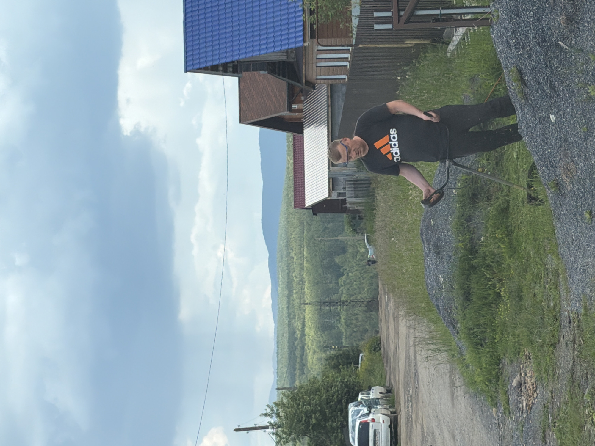 A man is trimming grass with a string trimmer in front of several houses in Mezhevoy, Russia. The man, wearing a black t-shirt with an orange Adidas logo and dark pants, is partially obscured by a pile of gravel. In the background, several cars are parked on a dirt road, and a line of trees stretches towards a distant mountain range under a cloudy sky. The houses have various colored roofs, including blue and brown.