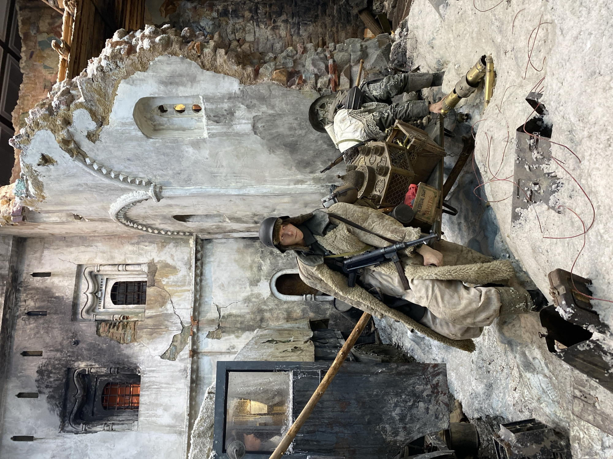 The image portrays a diorama of a scene depicting urban warfare, likely set in Moscow, Russia. The scene is staged within the ruins of a building with damaged walls and window frames. A mannequin dressed in a World War II-era Soviet winter uniform is holding a submachine gun. Behind it is a stove, and above, another soldier lies dead. There are also other historical military artifacts. The scene is highly detailed with realistic rubble, worn paint, and simulated damage, suggesting a museum exhibit.