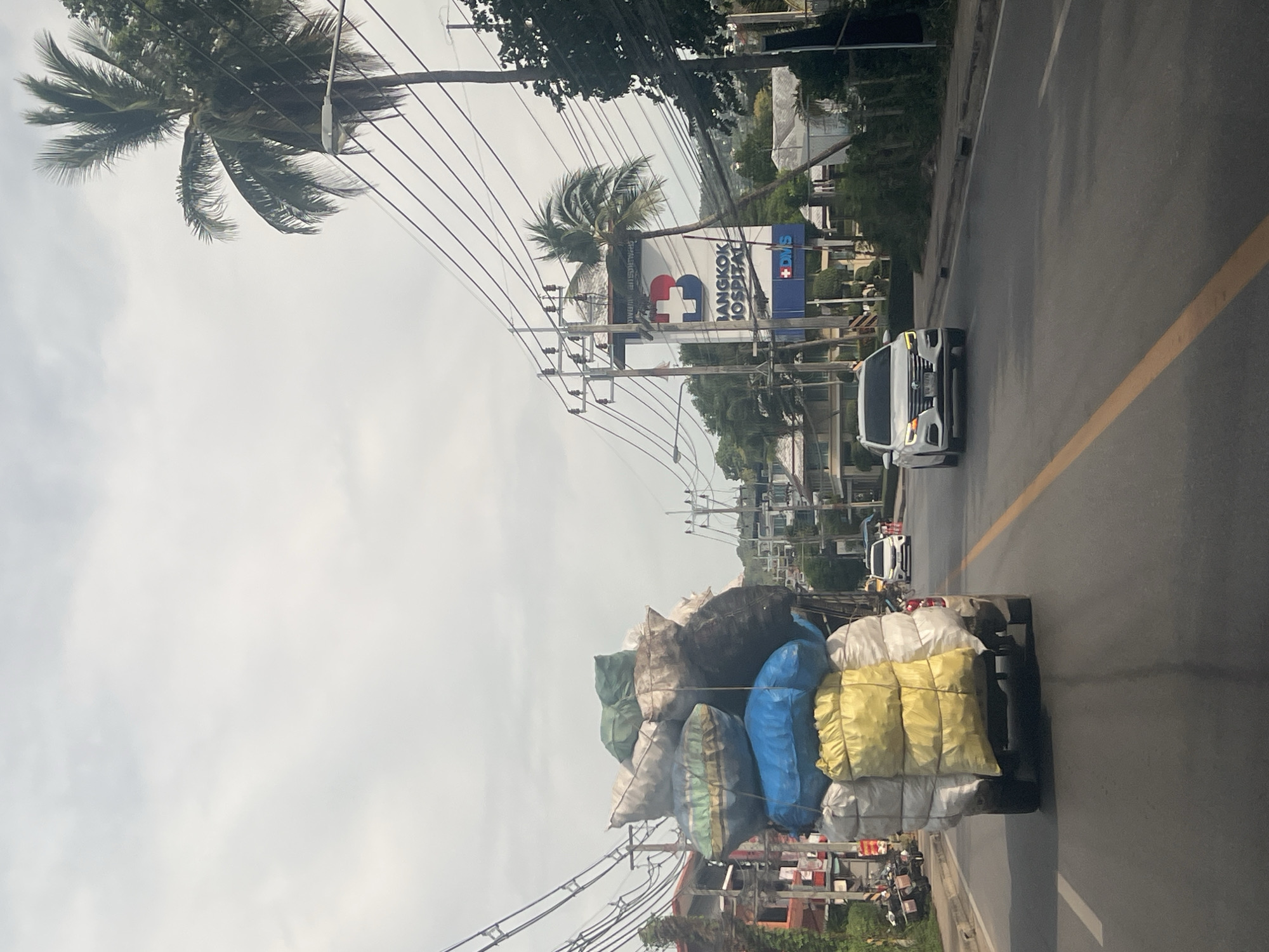The image depicts a street scene in Ko Samui District, Thailand, under an overcast sky. A heavily laden pickup truck, overflowing with large, colorful sacks of what appears to be goods, is traveling down a paved road. The sacks are stacked precariously high, with various colors including yellow, blue, green, white, and black. In the foreground, the rear of the pickup truck is visible.
To the right of the truck, a white sedan is driving in the same direction. Further down the road, a large billboard for "Bangkok Hospital" is prominently displayed, featuring a blue 'B' logo with a red cross. Palm trees and electrical wires are visible throughout the scene, typical of a tropical urban environment. Buildings and other vehicles are present on either side of the road, suggesting a moderately busy street. The overall impression is of everyday life and commerce in a Thai city.