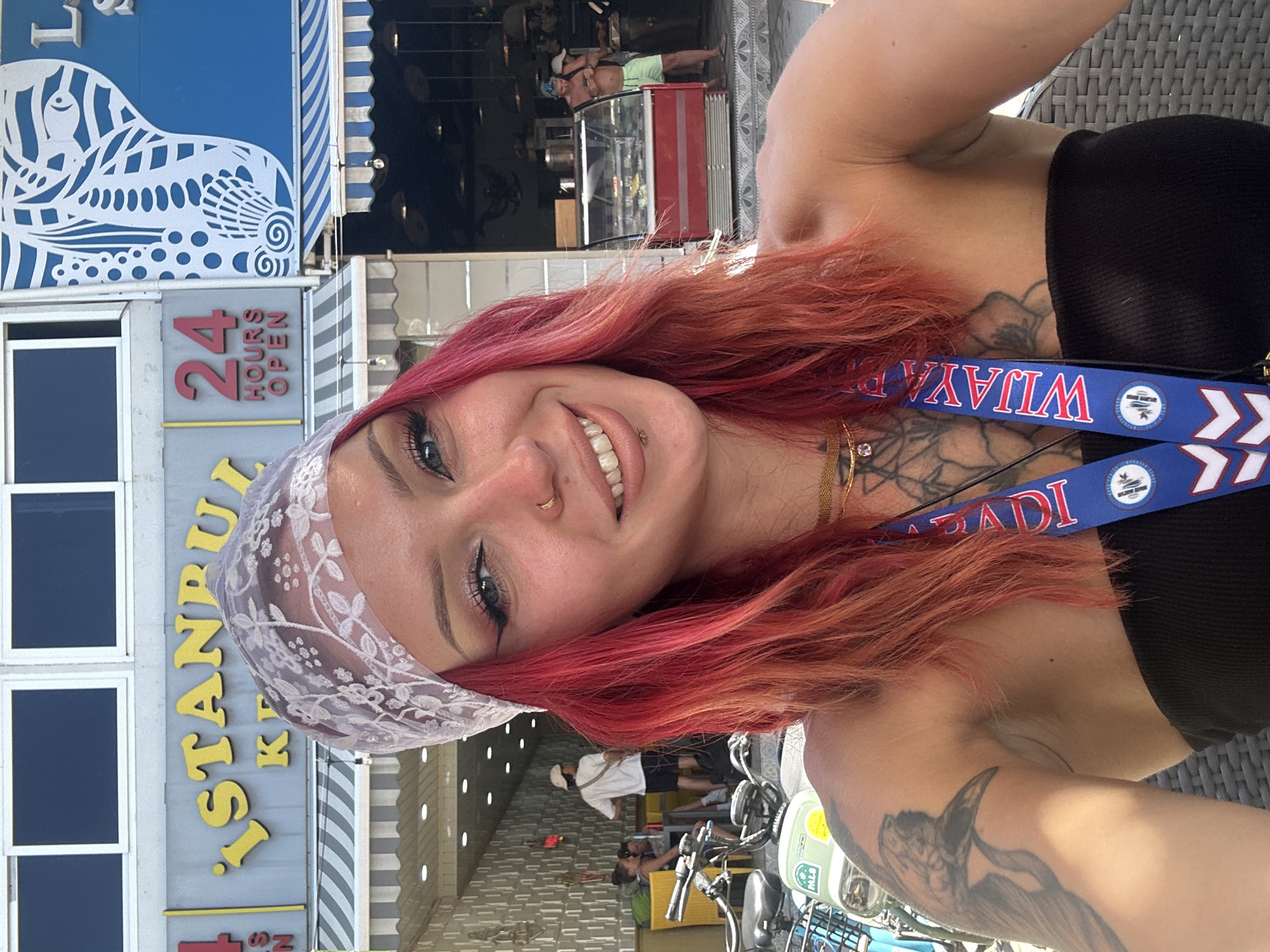 A selfie-style photograph shows a woman with red hair and fair skin, wearing a white lace headscarf and a black top. She has visible tattoos on her neck, chest, and arm. She is also wearing a lanyard around her neck.
Behind the woman, there is a sign for "ISTANBUL KOK". The sign also shows that the establishment is "24 HOURS OPEN" and a fish.
In the background, there are other people walking around. The location is Unknown, Indonesia. There are also parked bicycles visible. The photo appears to be taken outdoors during daylight.
