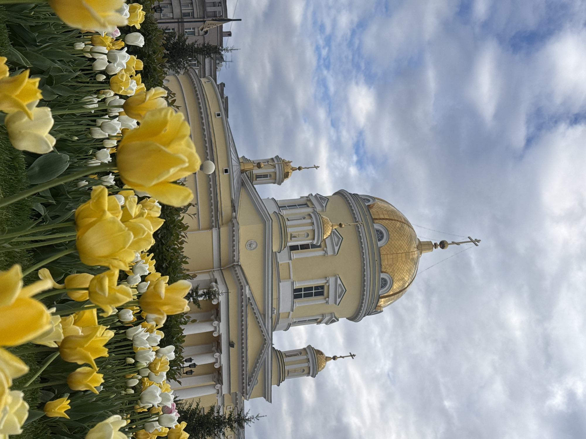 The image shows the Church on Blood in Yekaterinburg, Russia, from a low angle through a bed of yellow and white tulips. The church is pale yellow with white architectural detailing and a large, golden dome topped with a cross. The sky is cloudy with patches of blue. Buildings are visible in the background.